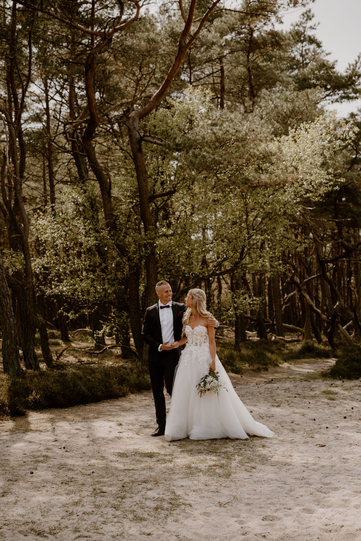 A couple in wedding attire stands together on a sandy path in a serene forest setting.