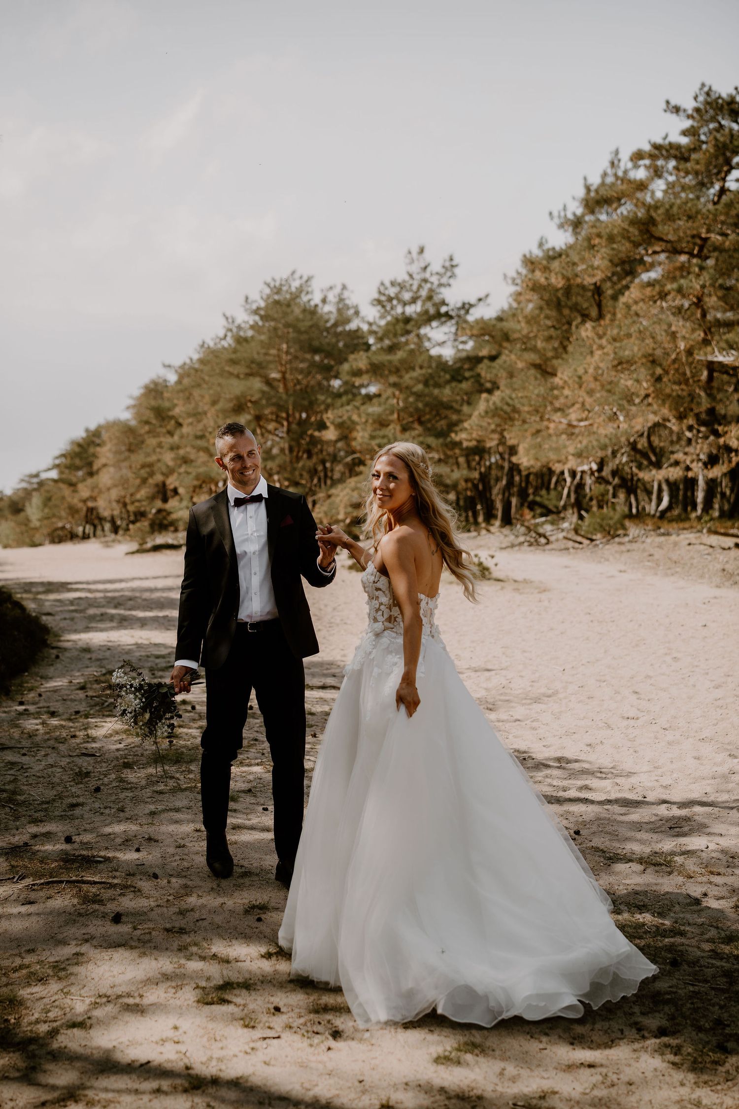 A couple on their wedding day walking along a sandy forest path with pine trees in the background.
