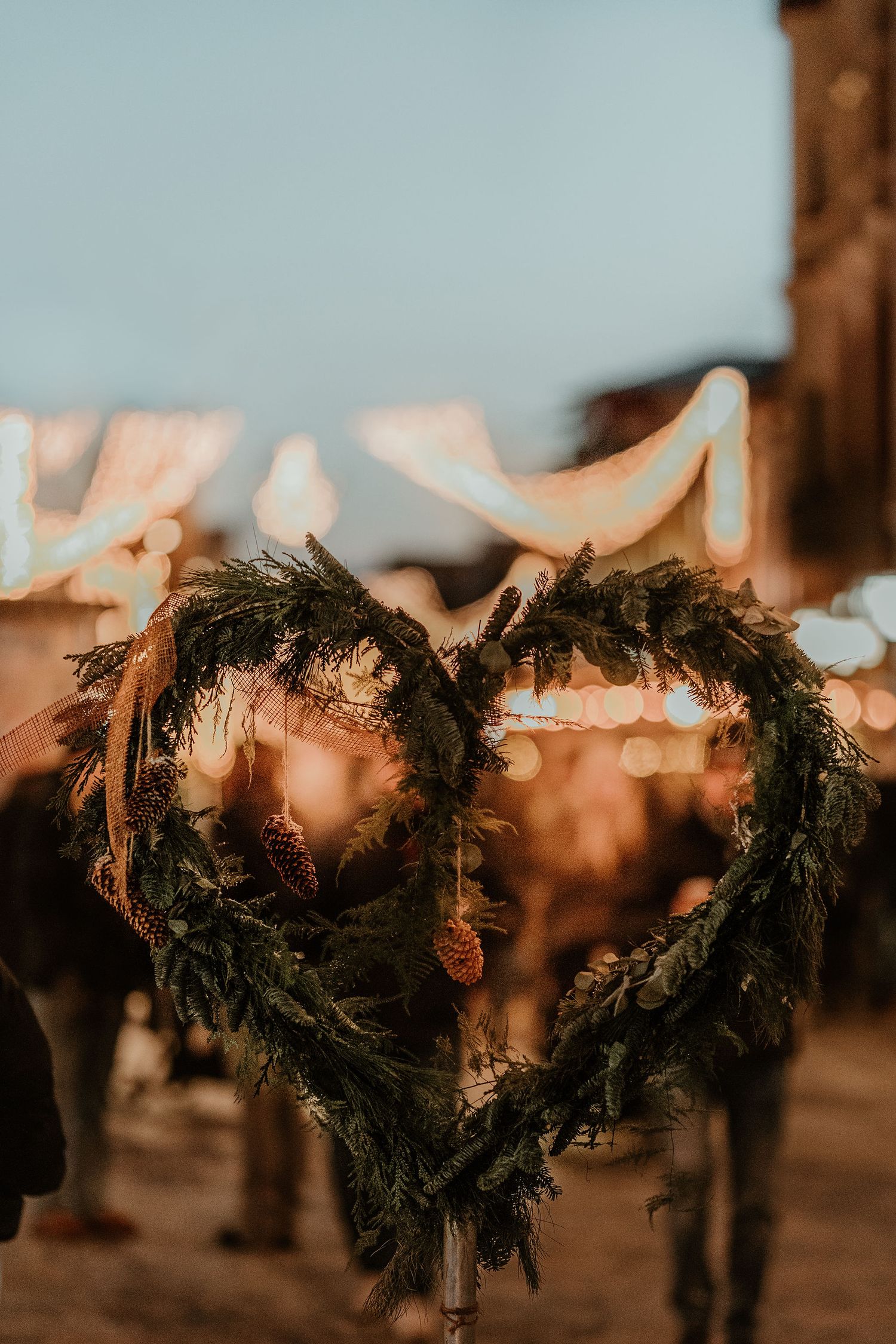 A rustic heart-shaped wreath made of dark evergreen branches illuminated by warm string lights in a romantic setting.