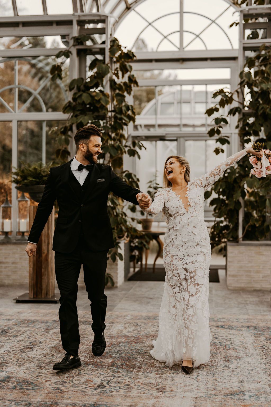 A joyful couple laughs together in a greenhouse setting wearing wedding attire, surrounded by lush greenery and arched windows.