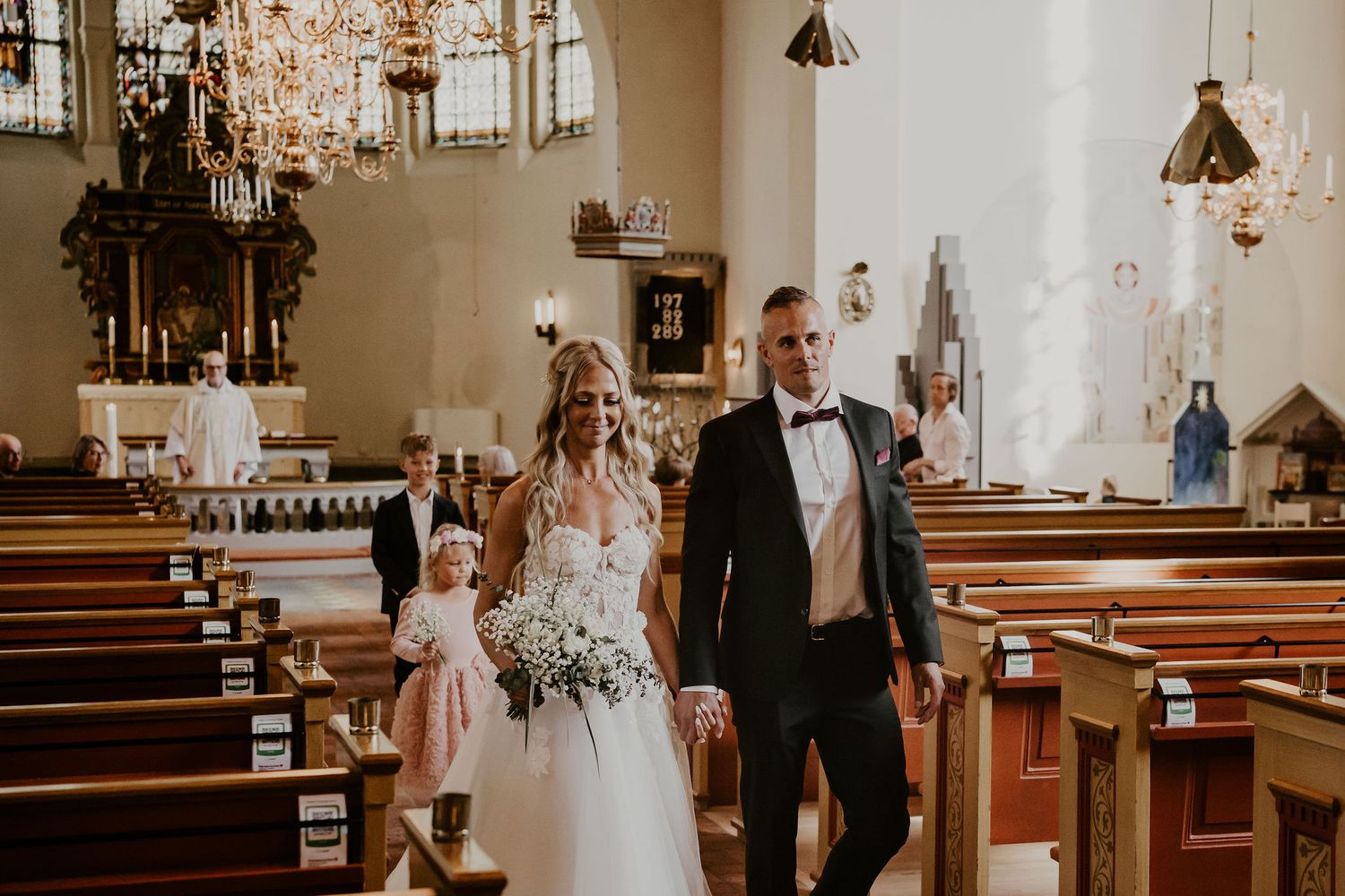 Newlyweds walking down the aisle in a traditional church with elegant chandeliers and wooden pews.
