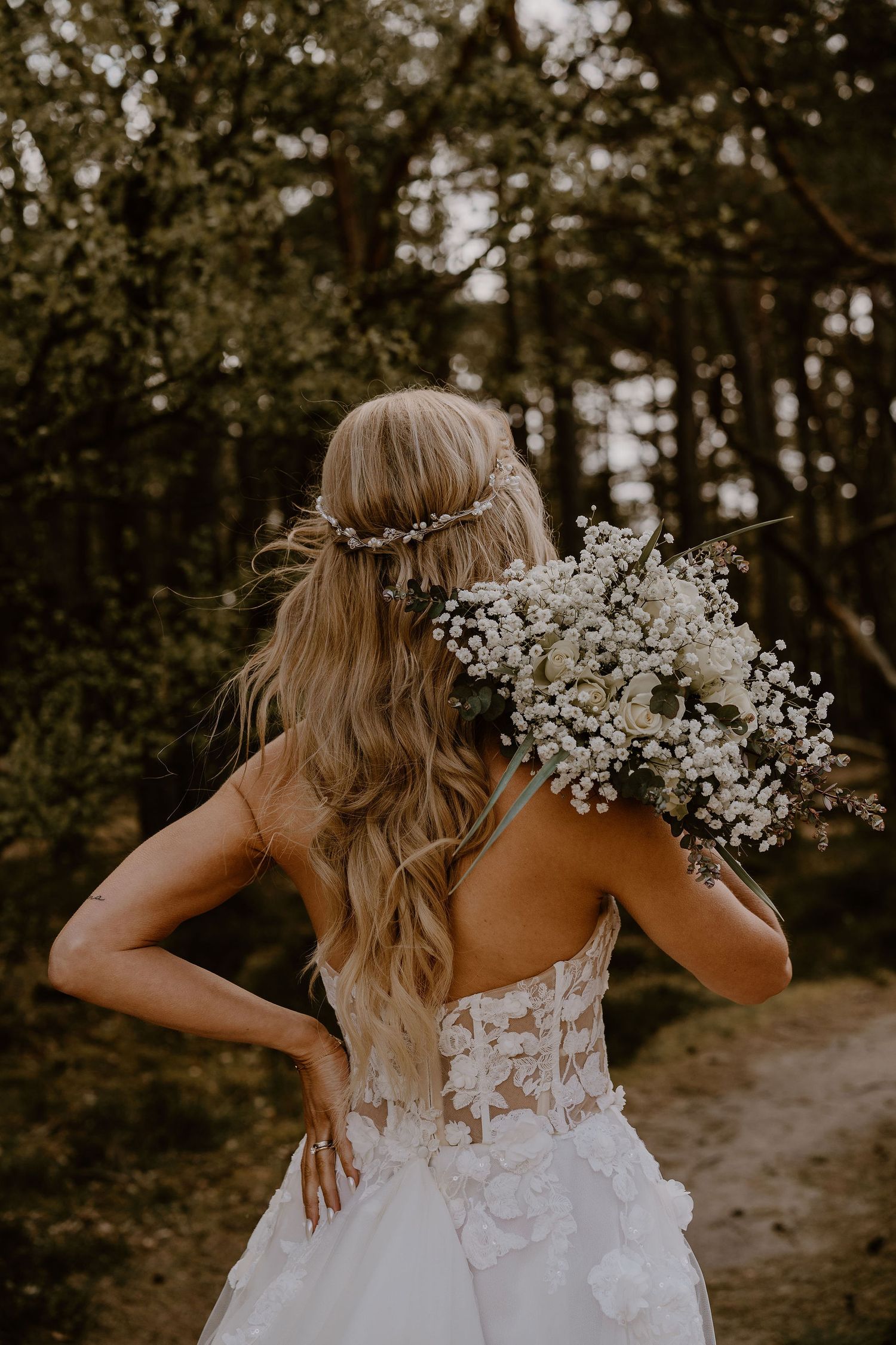 Back view of a bride in a white lace dress holding baby's breath flowers in a forest setting at sunset.