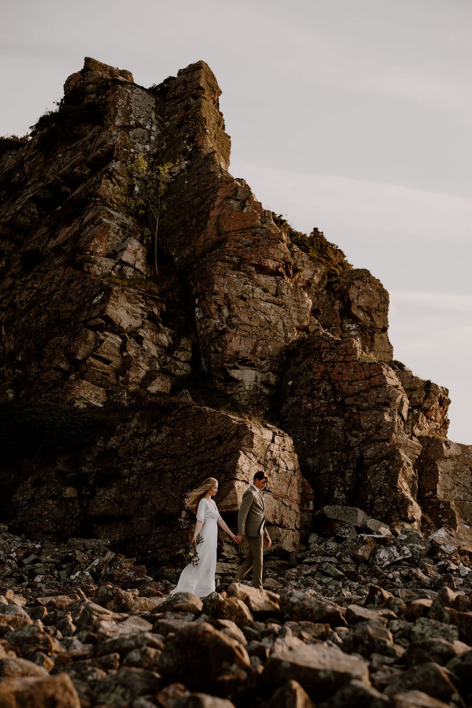 Two people stand together on rocky terrain beneath towering cliffs at sunset.