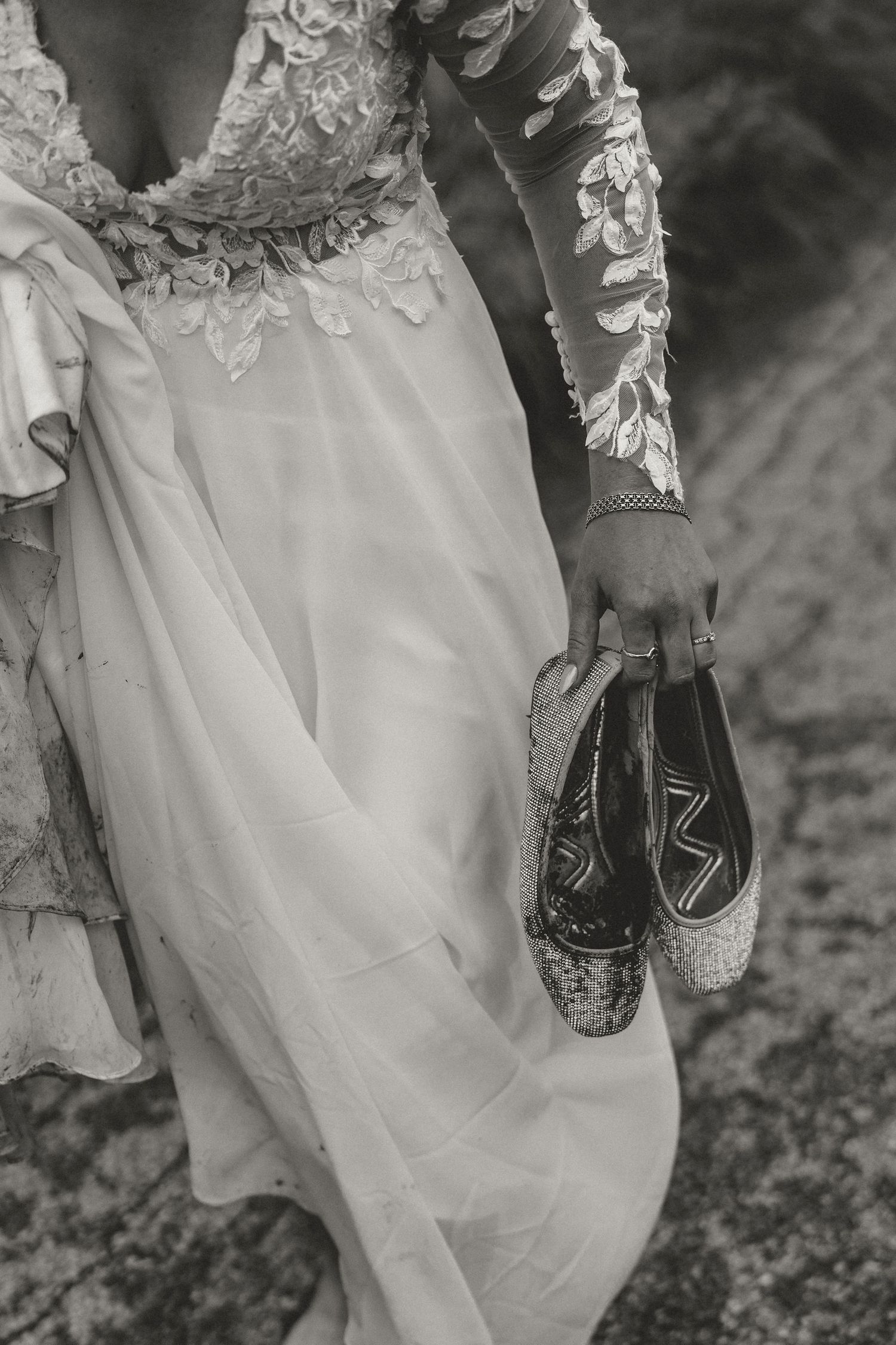 Wedding dress detail showing intricate lace sleeves and sparkly bridal shoes held against flowing white fabric.