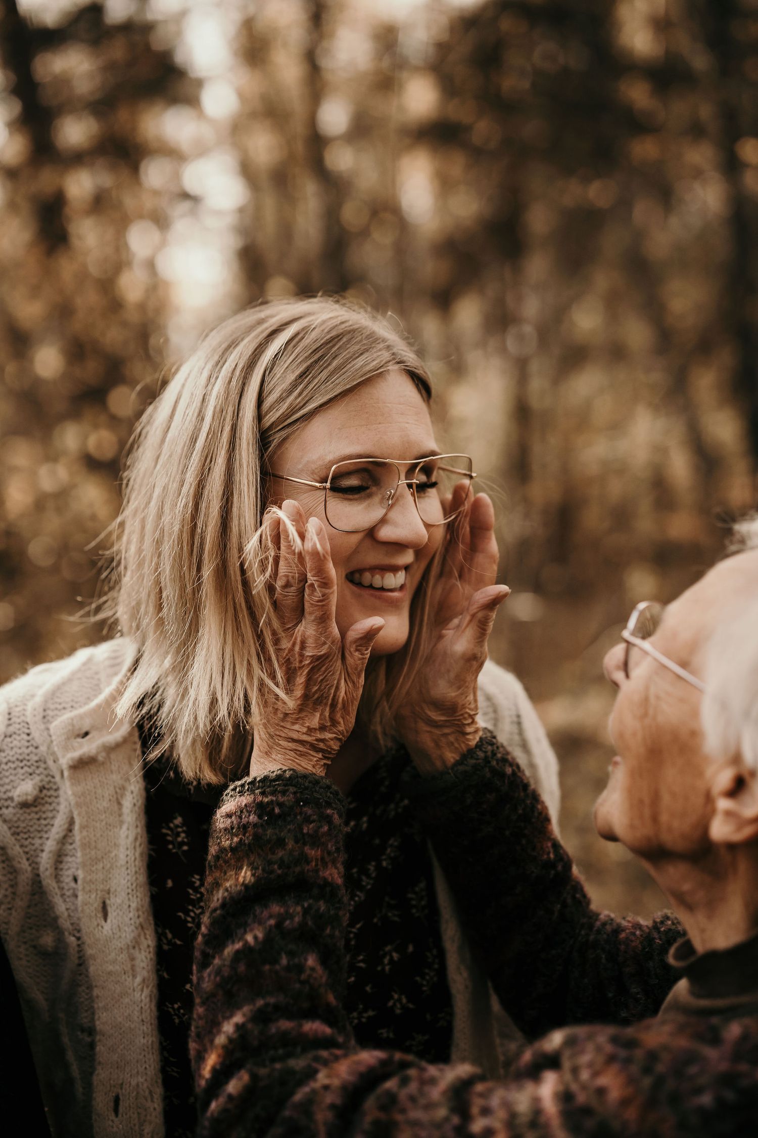 Elderly person in dark sweater touching face of younger person wearing glasses in sepia-toned outdoor setting.