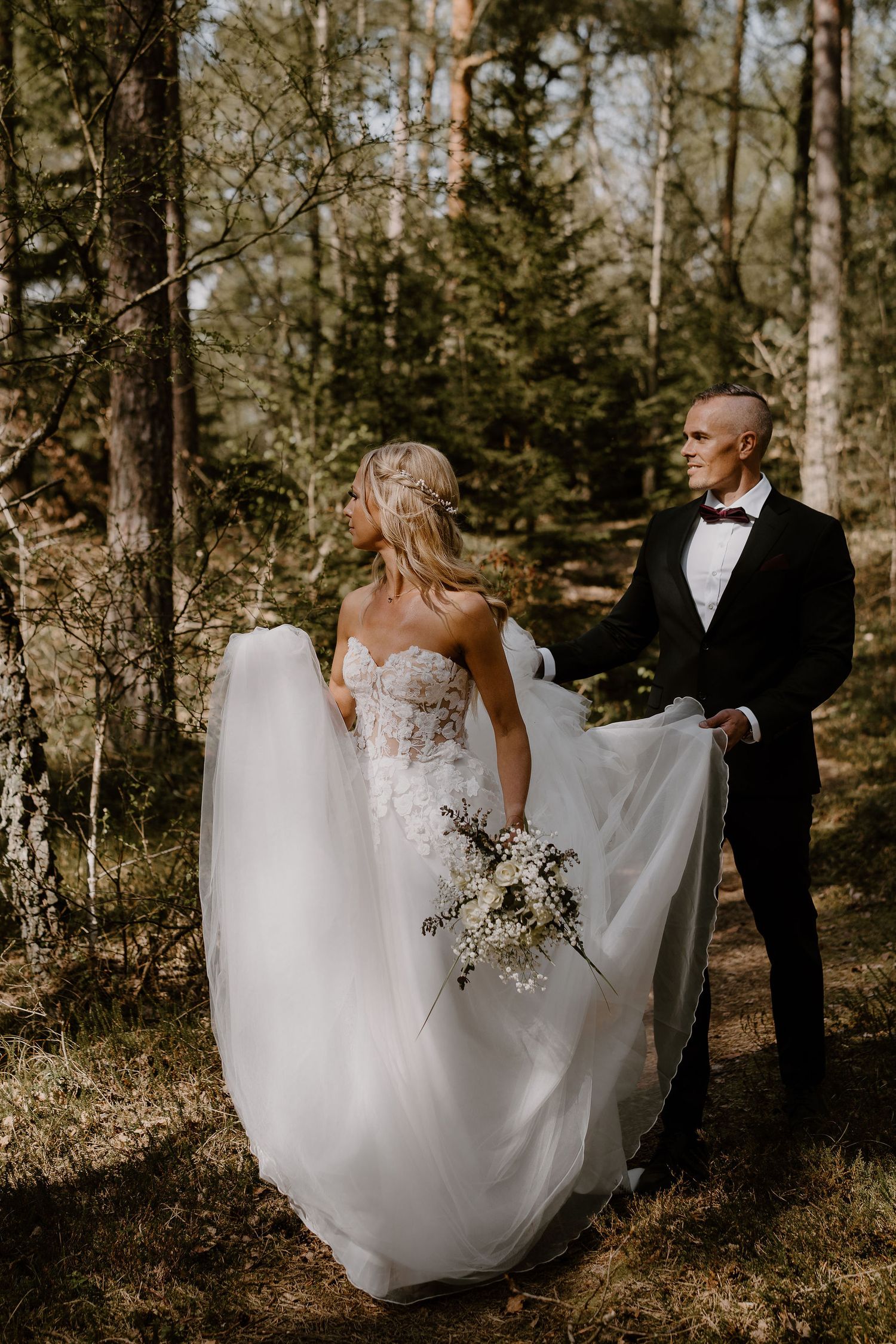 Couple in formal wedding attire stands together in a sun-dappled forest setting with flowing white dress and bouquet.