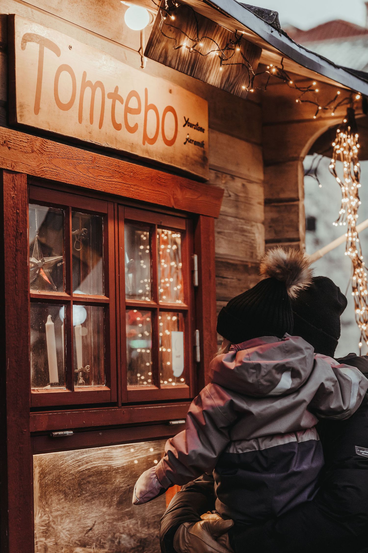 A cozy wooden storefront with 'Tomtebo' sign and twinkling string lights illuminating the window display on a winter evening.