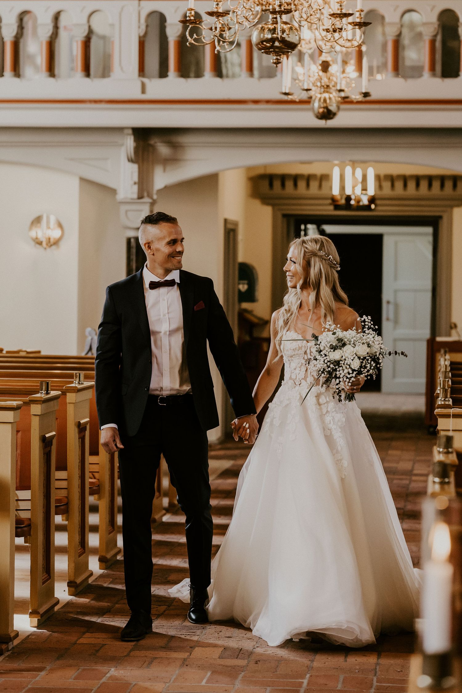 A wedding couple shares a joyful moment while walking down the church aisle among wooden pews and elegant chandeliers.