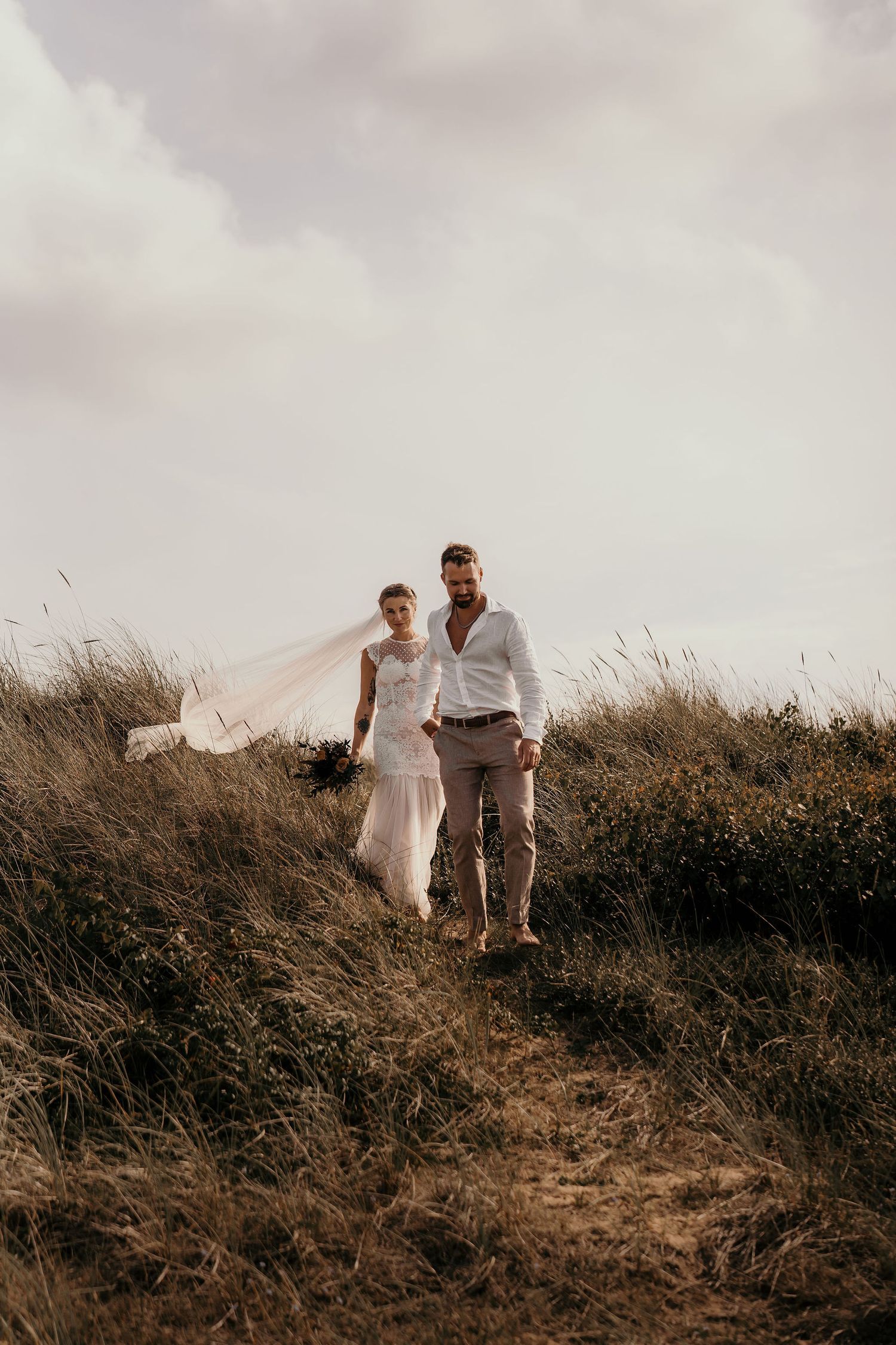 A couple in wedding attire walks together on a hillside path through tall grass at sunset.