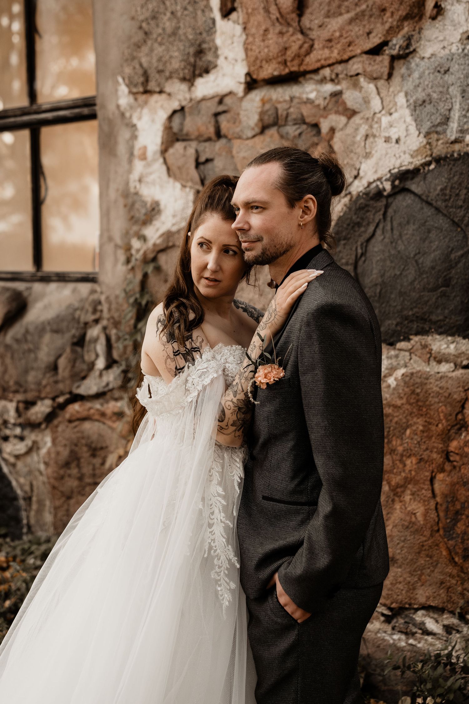 A couple poses against a rustic stone wall, wearing elegant wedding attire including a lace white dress and dark suit.