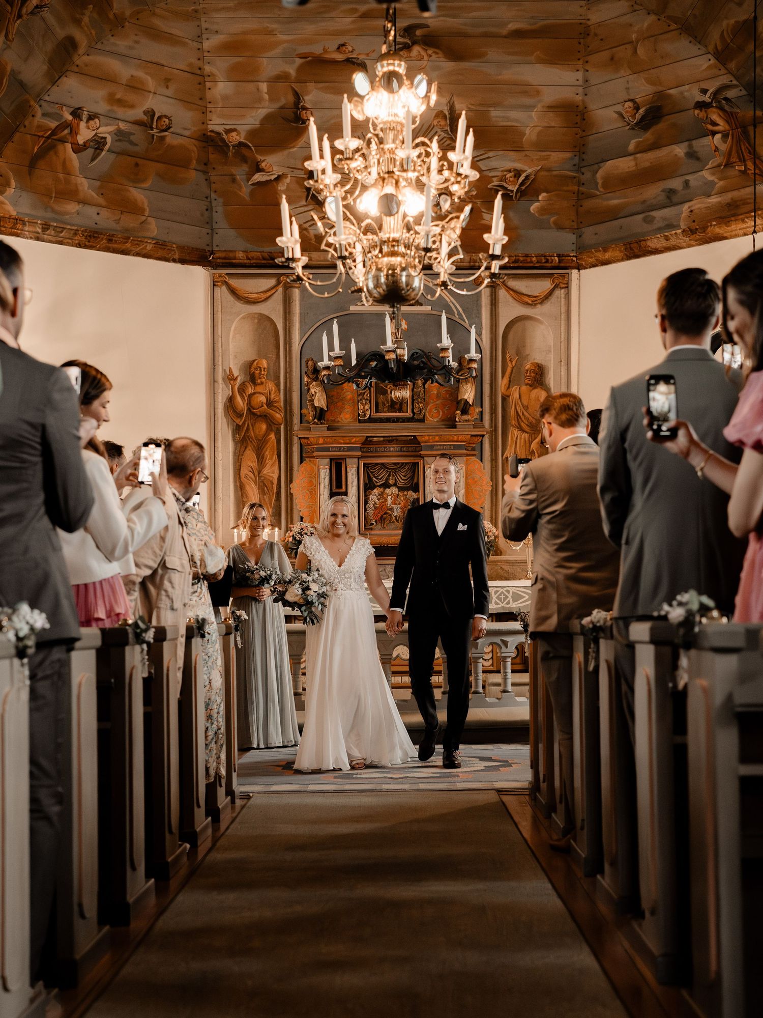 A wedding ceremony takes place in a historic church with a grand chandelier and wooden interior glowing in warm light.