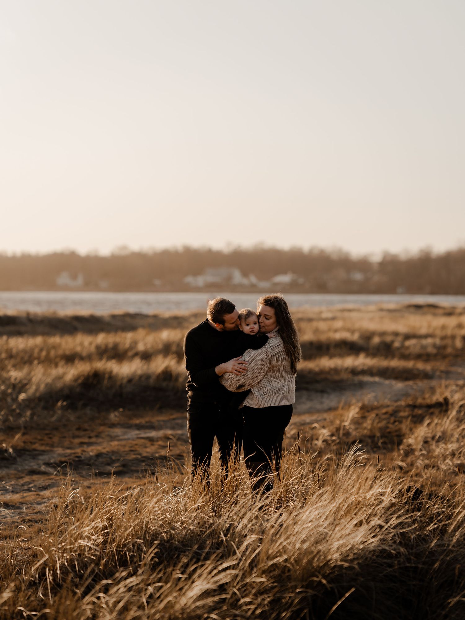 A couple embraces in tall golden grass during sunset with water and houses visible in the background.
