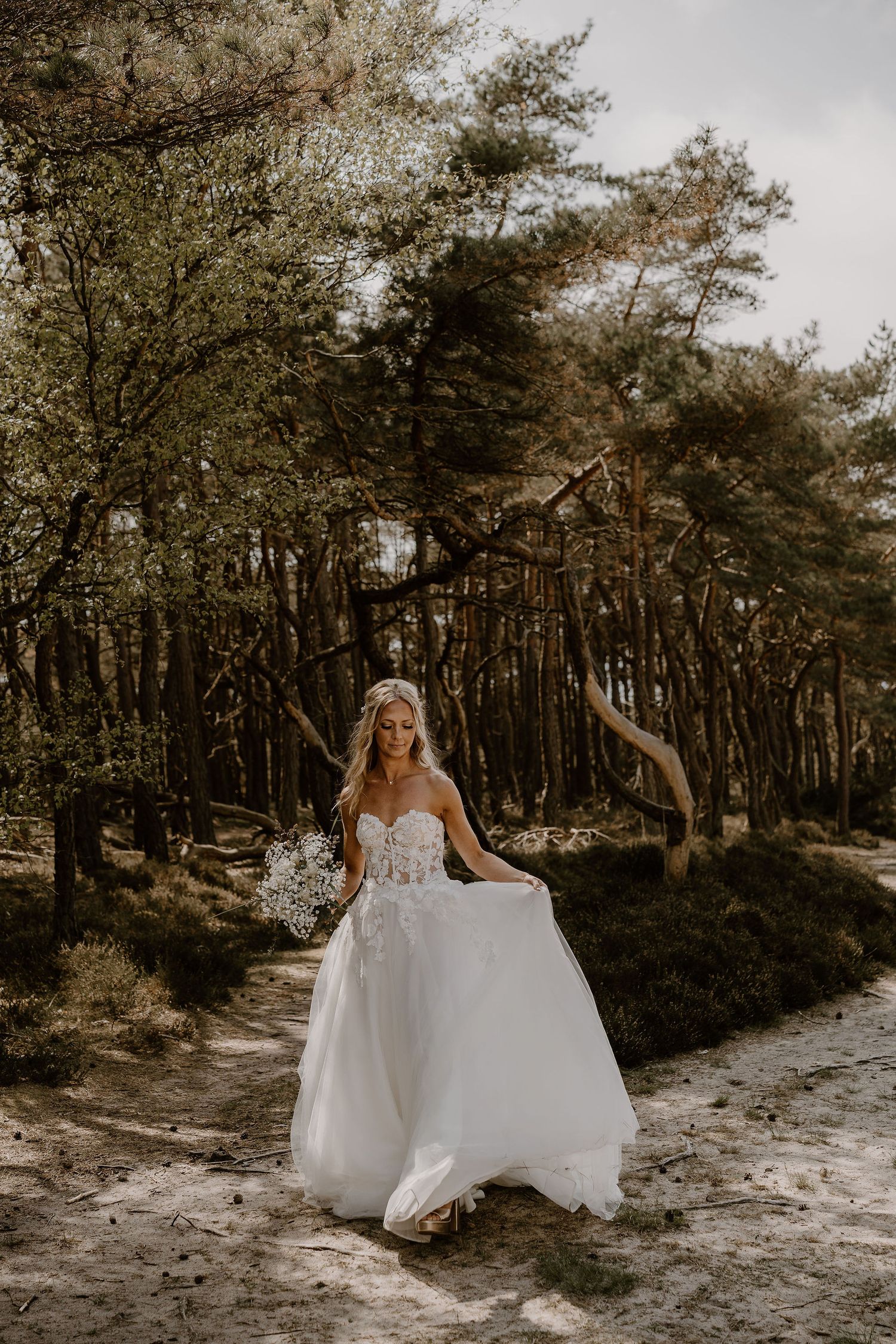 A person in an elegant white wedding dress poses gracefully on a sandy forest path surrounded by tall pine trees.