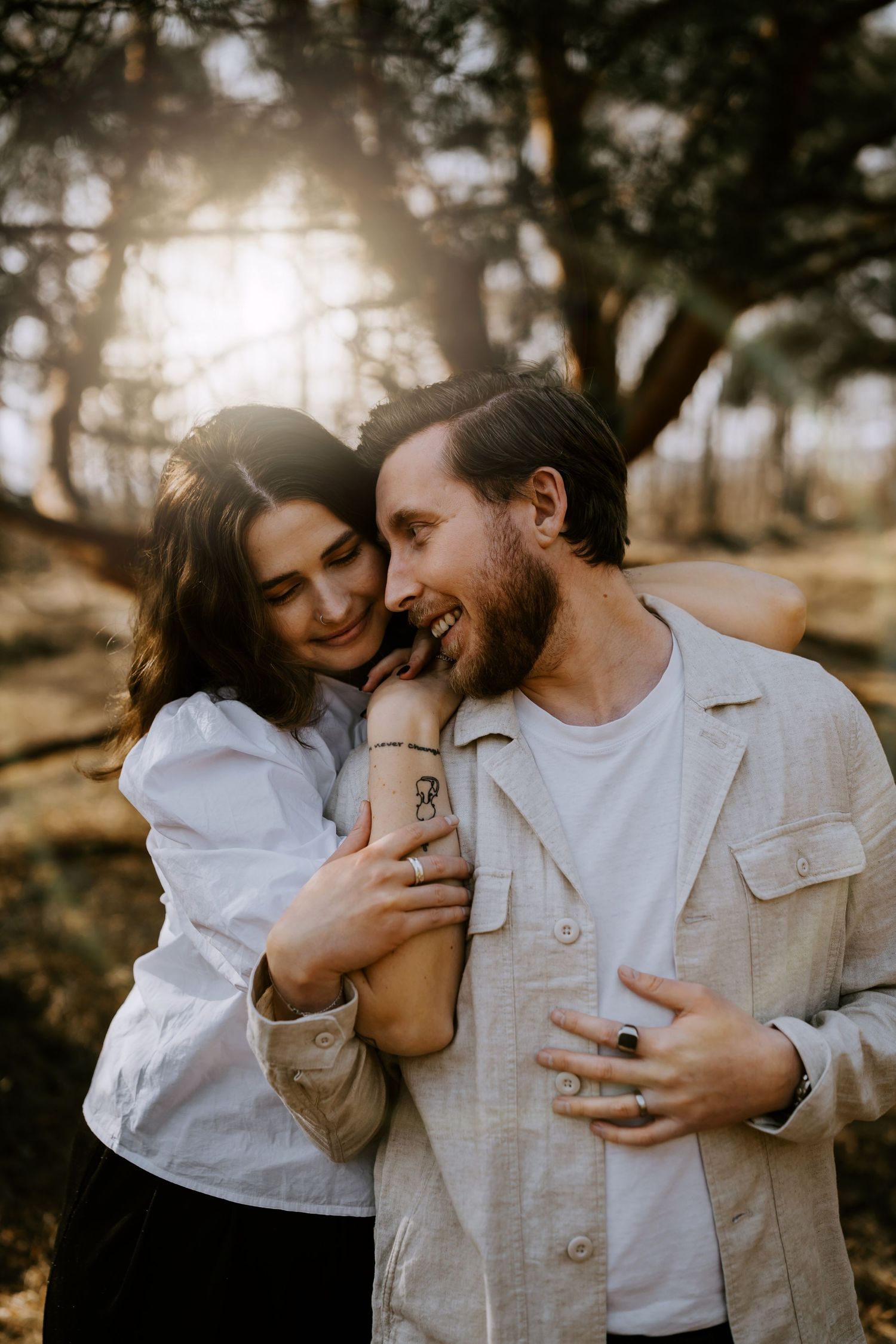 A romantic couple embraces and laughs together in a sunlit forest setting during golden hour.