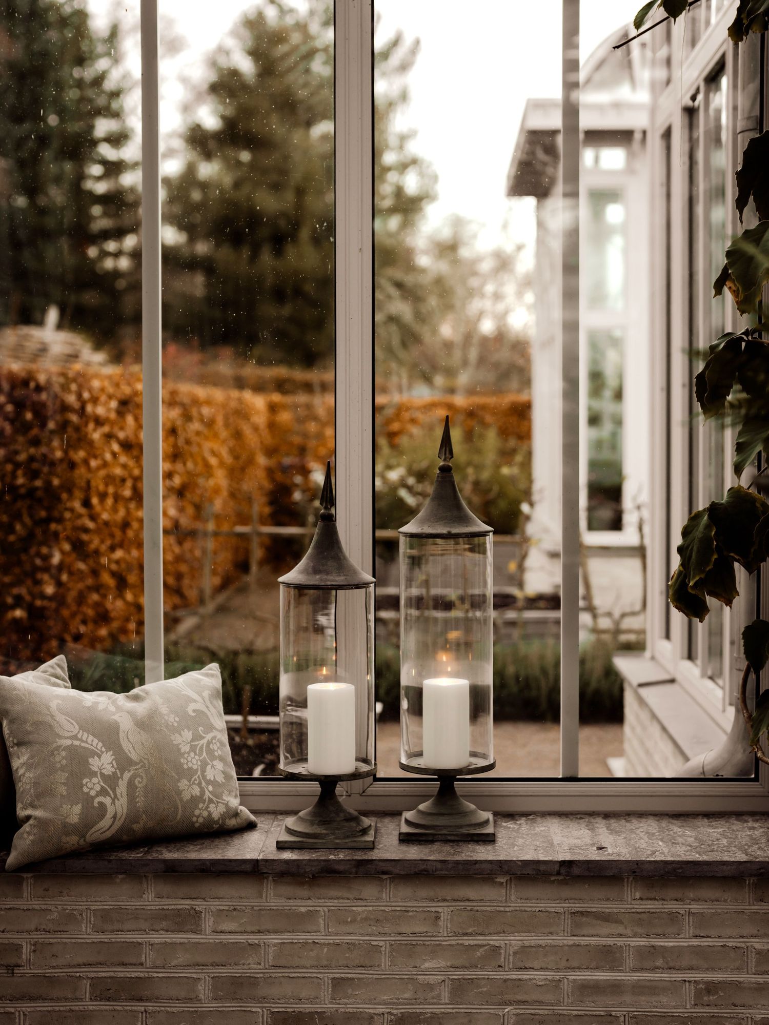 Cozy window nook with lanterns and decorative pillows overlooking an autumn garden with hedges.