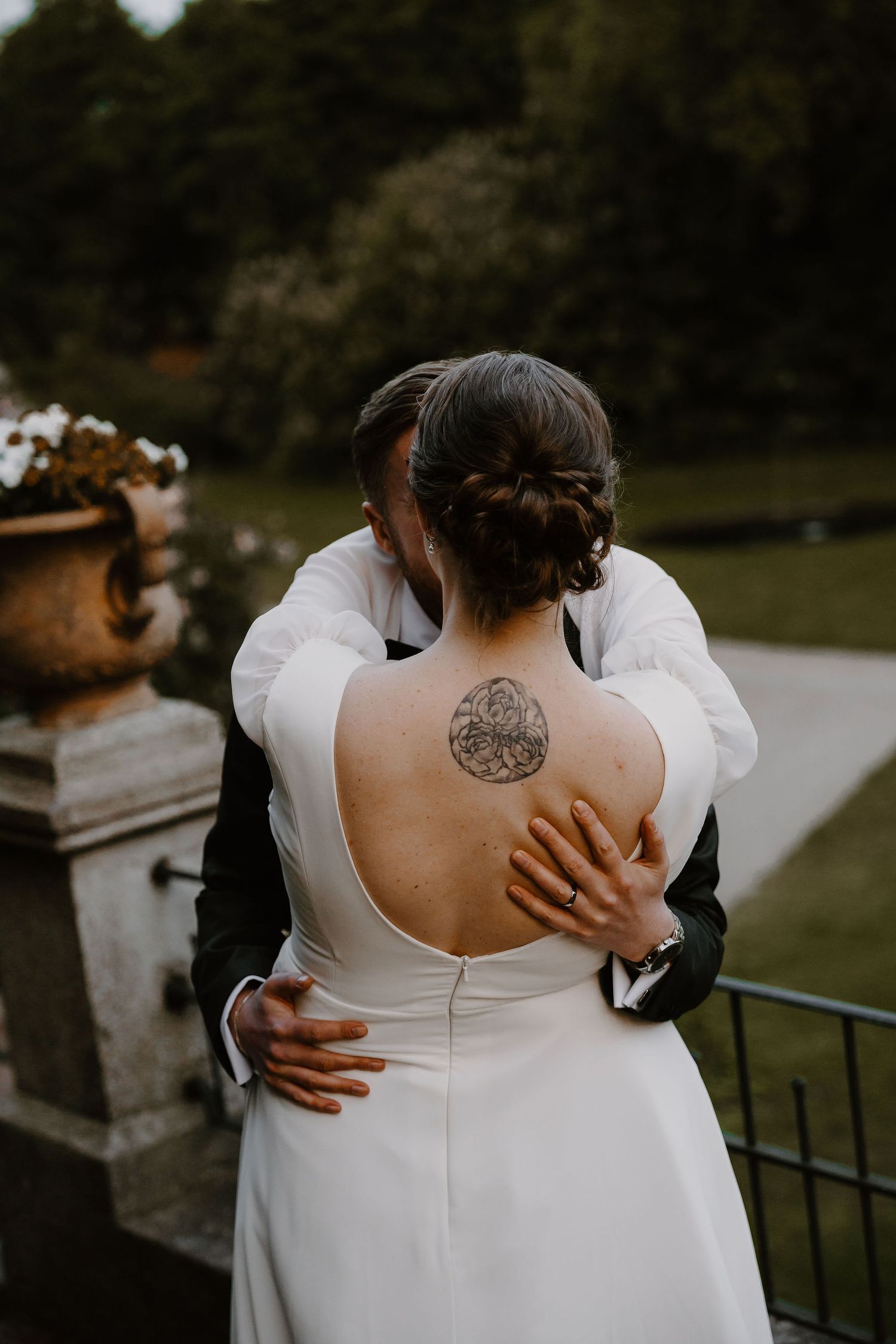 A bride in a white backless dress with a circular tattoo embraces someone during an outdoor wedding ceremony.