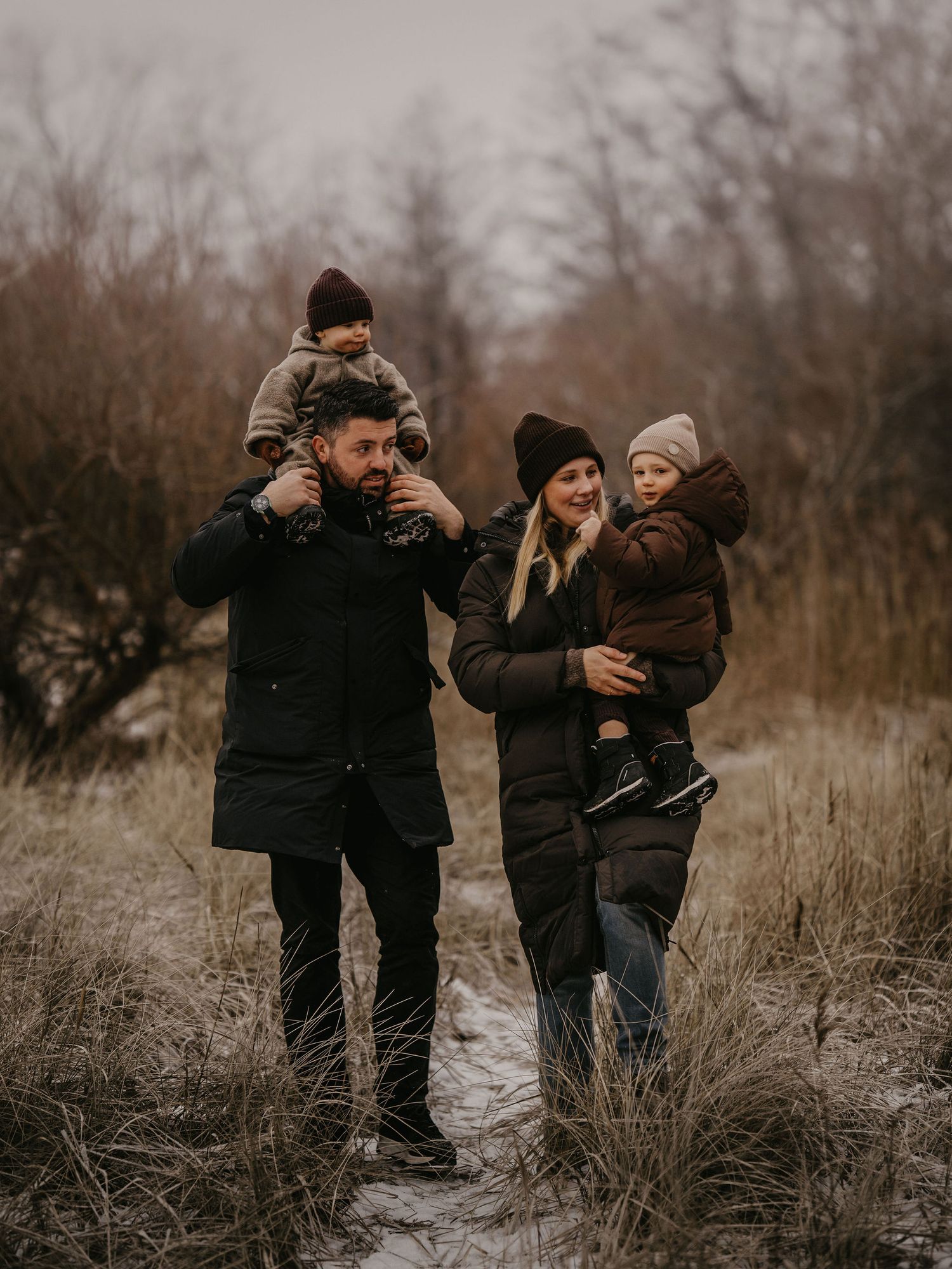 Family of four walking together through tall grass on a cold winter day in dark coats and warm hats.