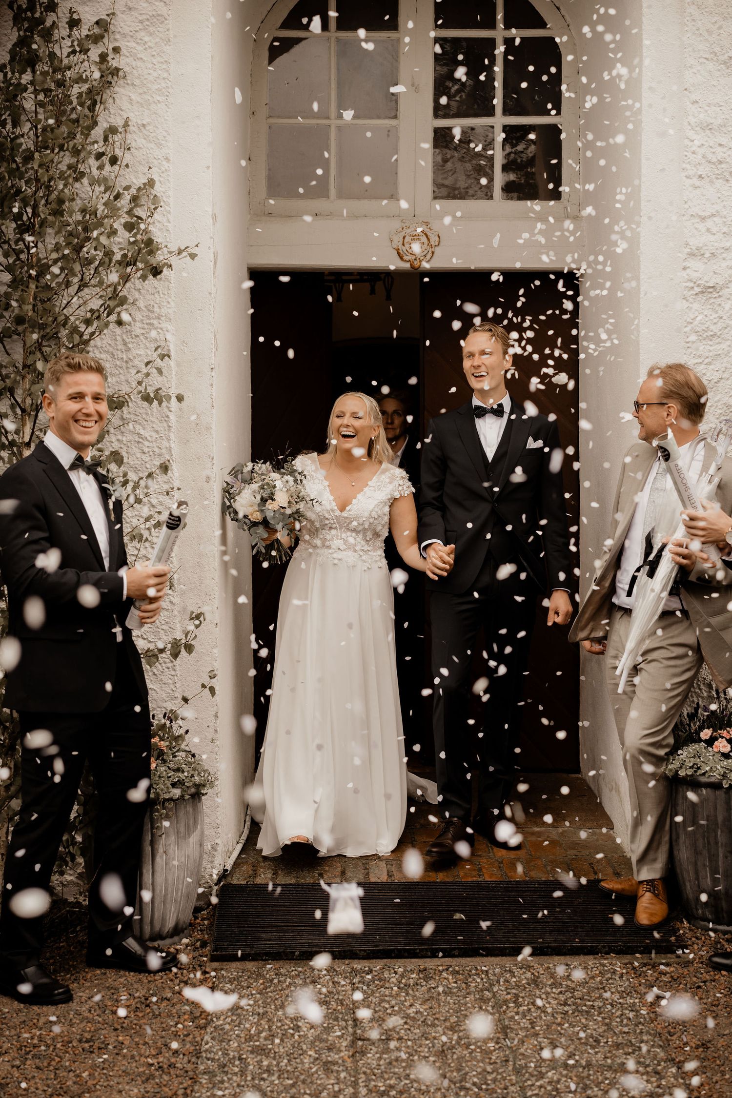 Wedding celebration at an elegant entrance with petals falling while joyful couple and guests share laughs on the steps.