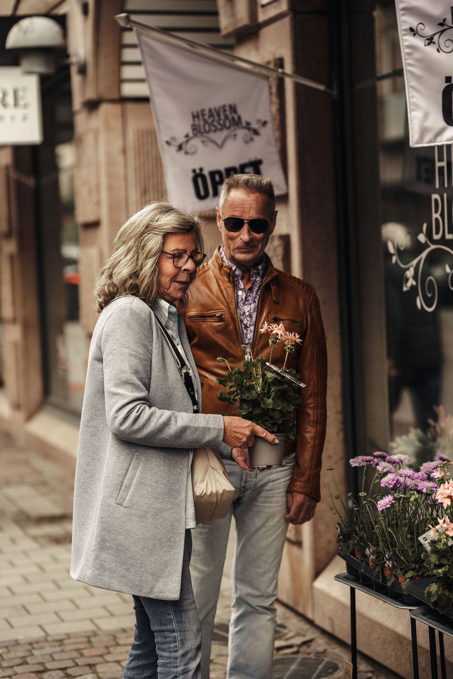 A stylishly dressed couple shops for flowers on a cobblestone street outside a market.