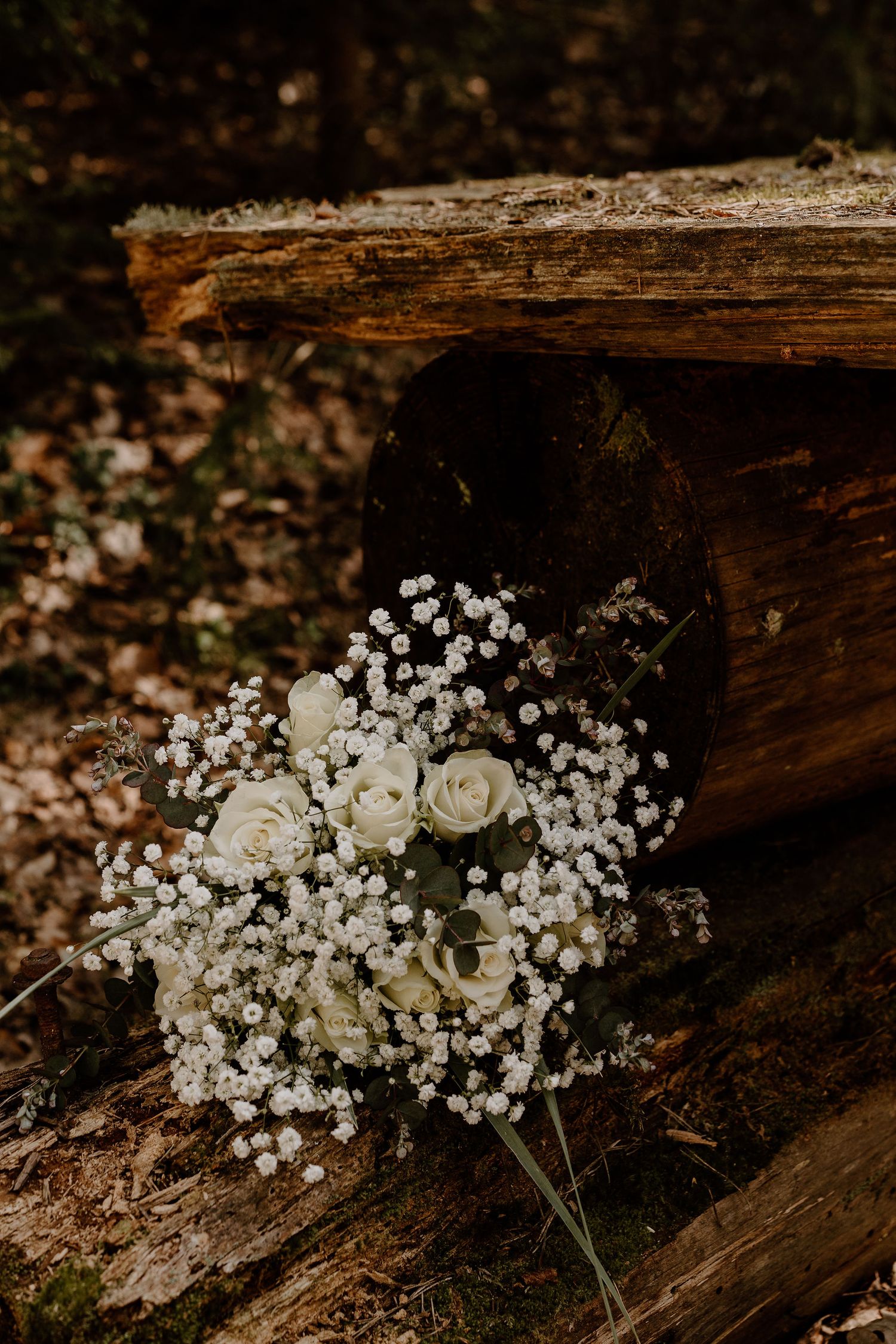 A delicate bridal bouquet with white roses and baby's breath rests against rustic wooden logs in moody lighting.