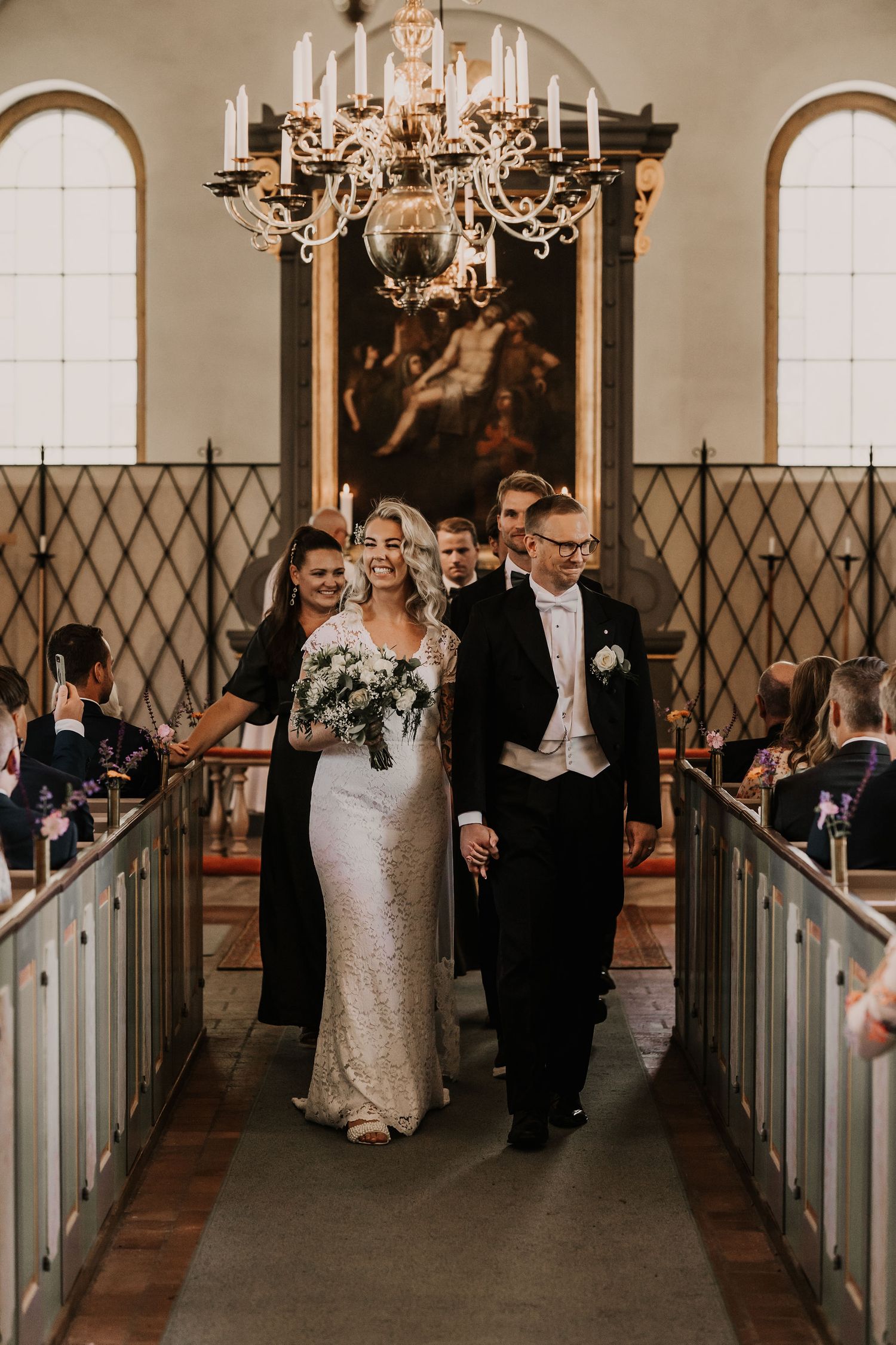 A wedding couple walks down the aisle of a historic church with elegant chandelier and arched windows.