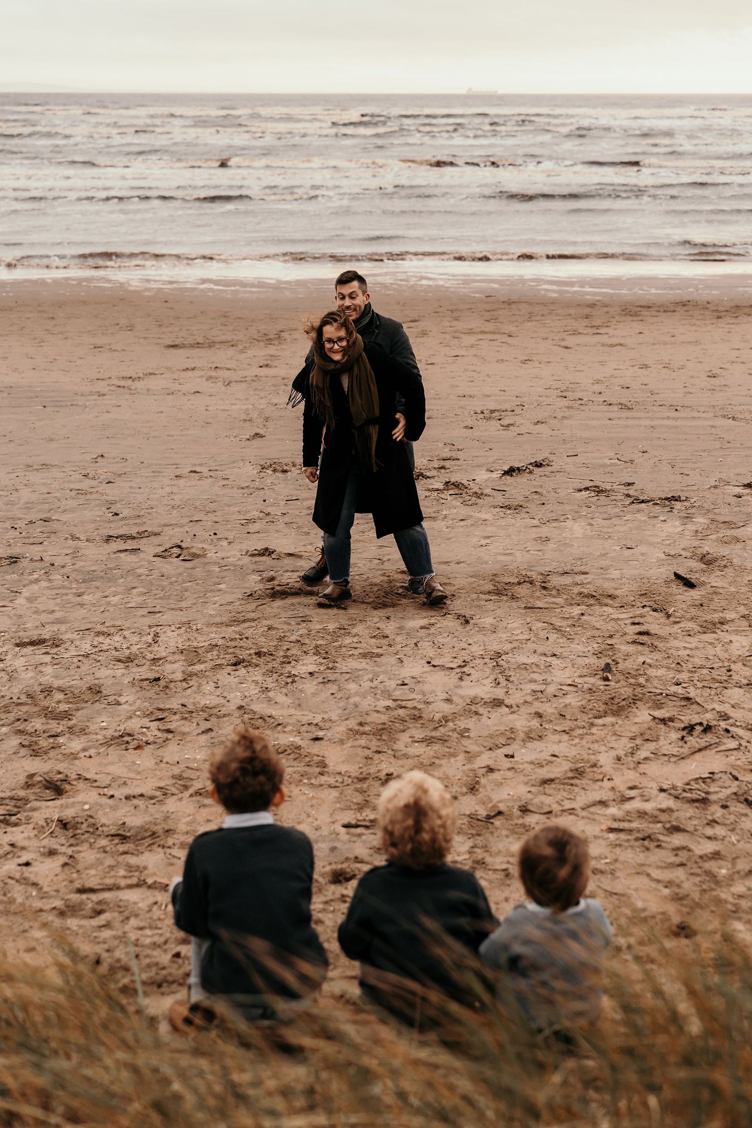 Three people sit on a grassy dune overlooking two figures standing on a misty beach at dusk.