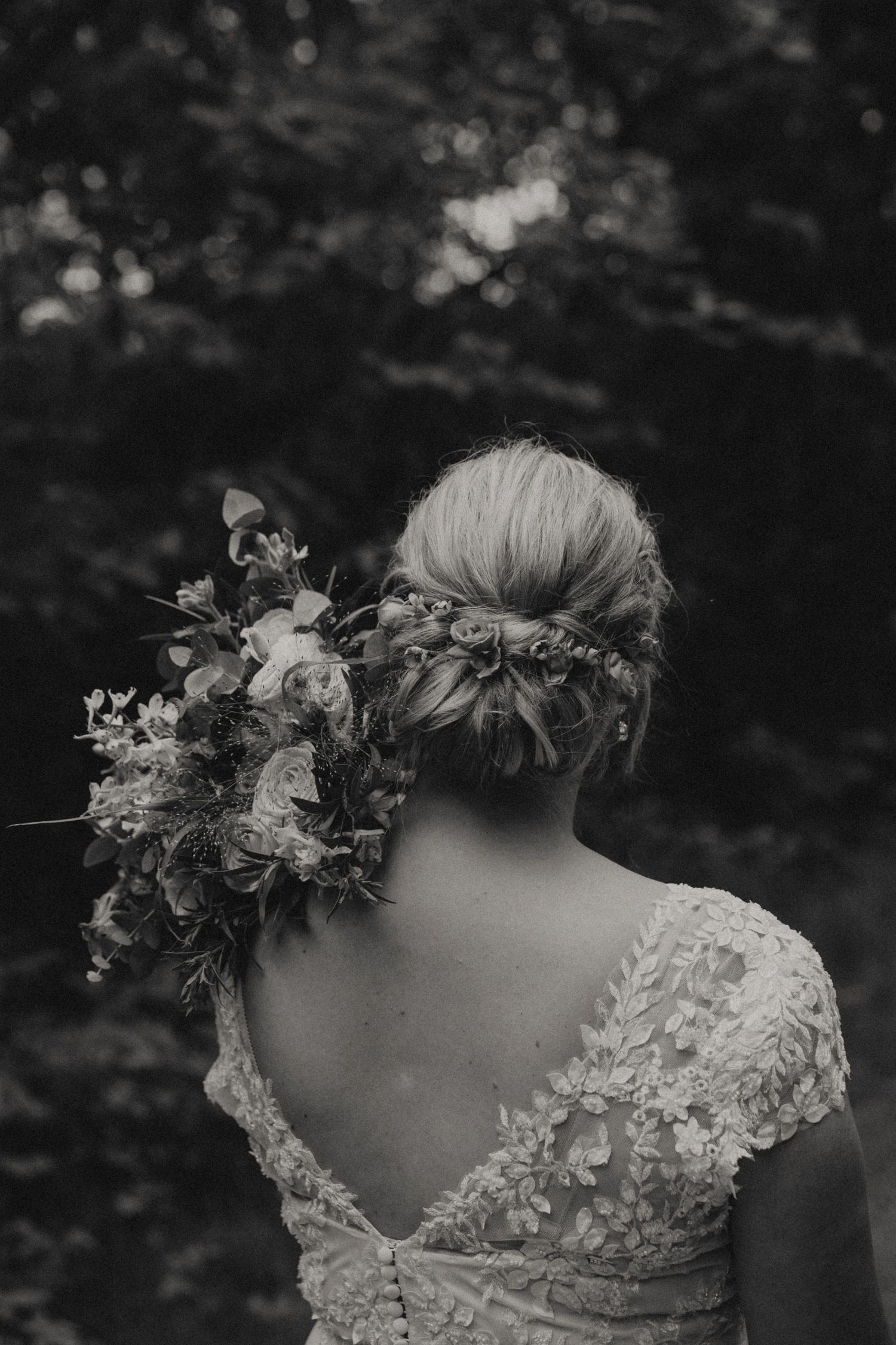 Black and white photo of a bride wearing a lace dress and floral hairpiece, shot from behind in a garden setting.