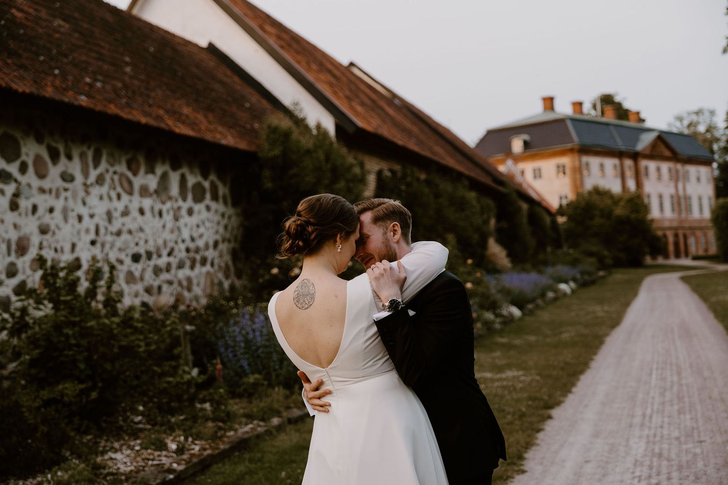 A romantic couple in wedding attire embrace on a stone path leading to a rustic building with white walls.
