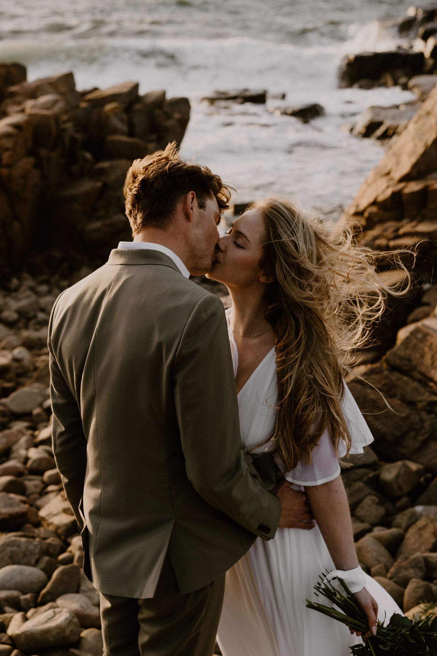 A romantic coastal wedding moment captured on rocky shore with windswept hair and intimate kiss at sunset.