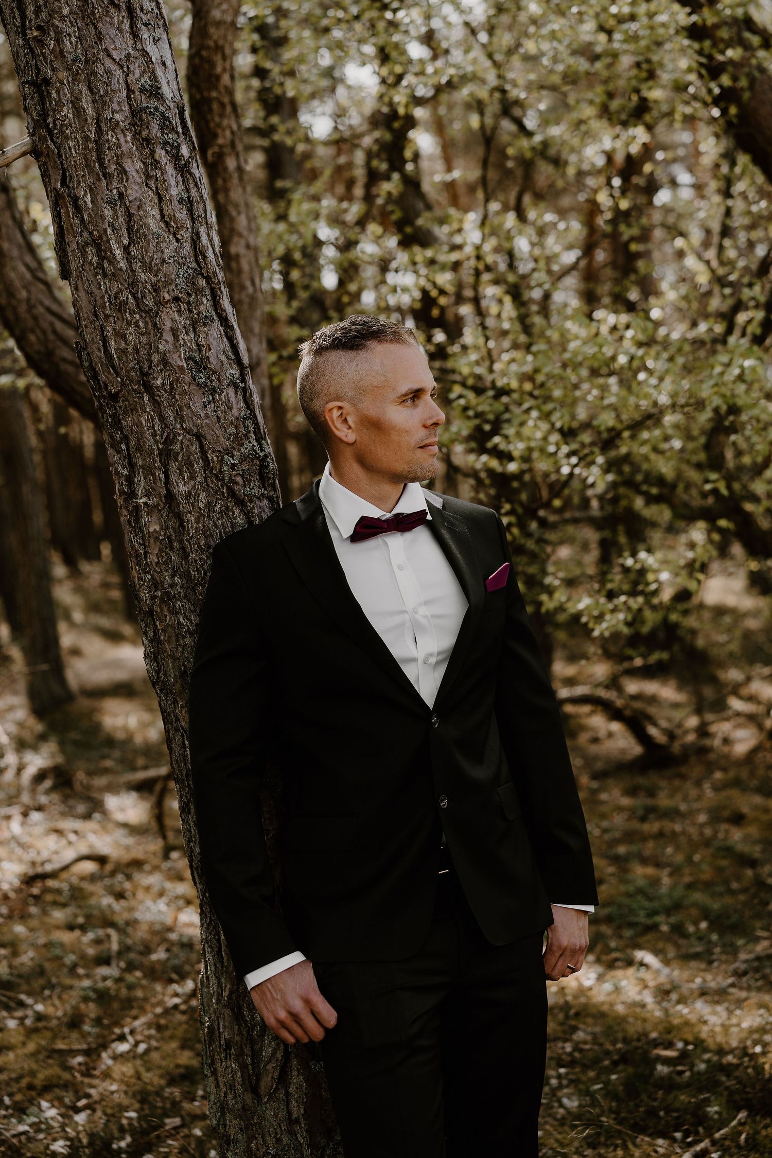 A person in a formal black tuxedo and bow tie poses against a tree trunk in a sunlit wooded setting.