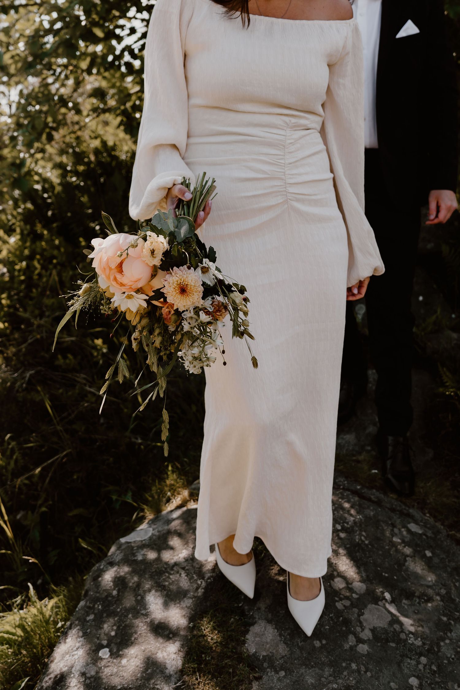 Modern wedding dress with long sleeves and square neckline shown with peach and white floral bouquet in sunlit garden setting.