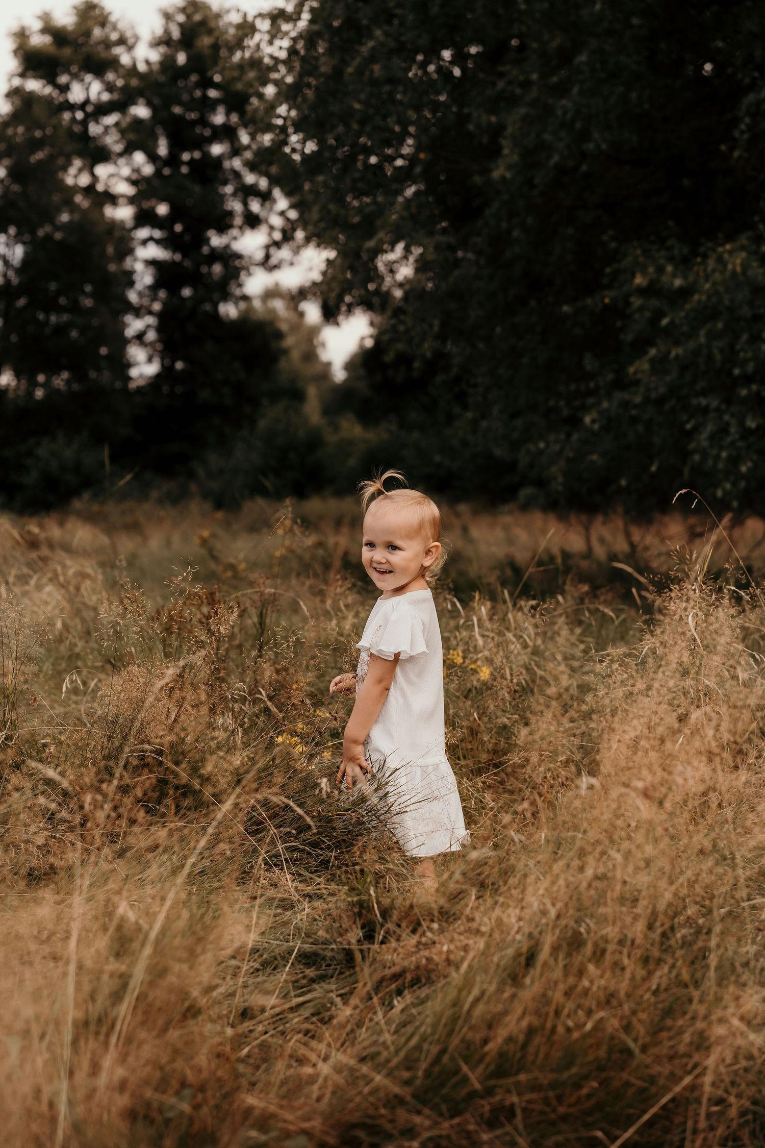Young child in white clothing standing in tall golden grass at sunset.