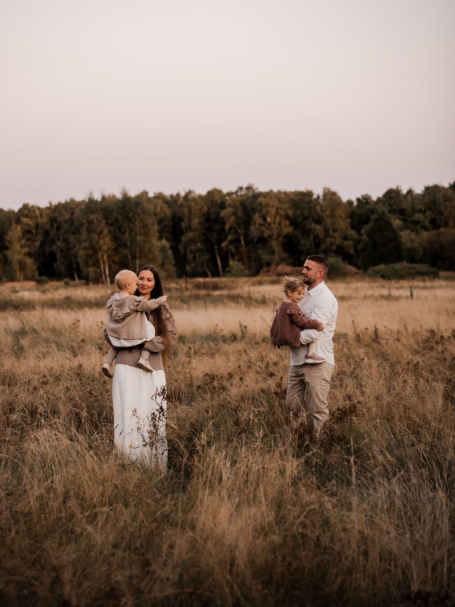 Two adults stand together in a golden autumn field during sunset while holding small children.
