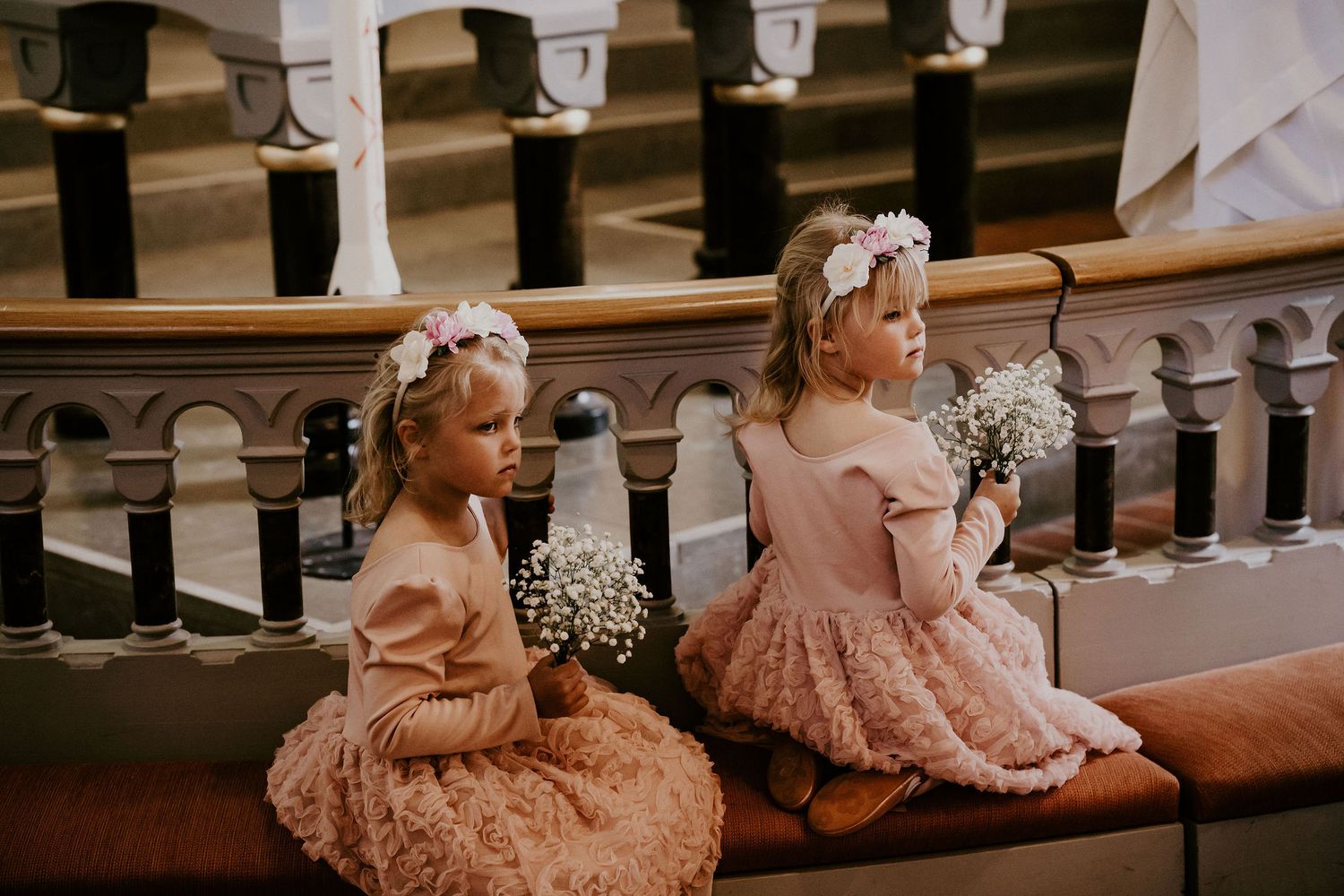 Two flower girls in pink dresses sit on wooden church pews holding baby's breath bouquets.