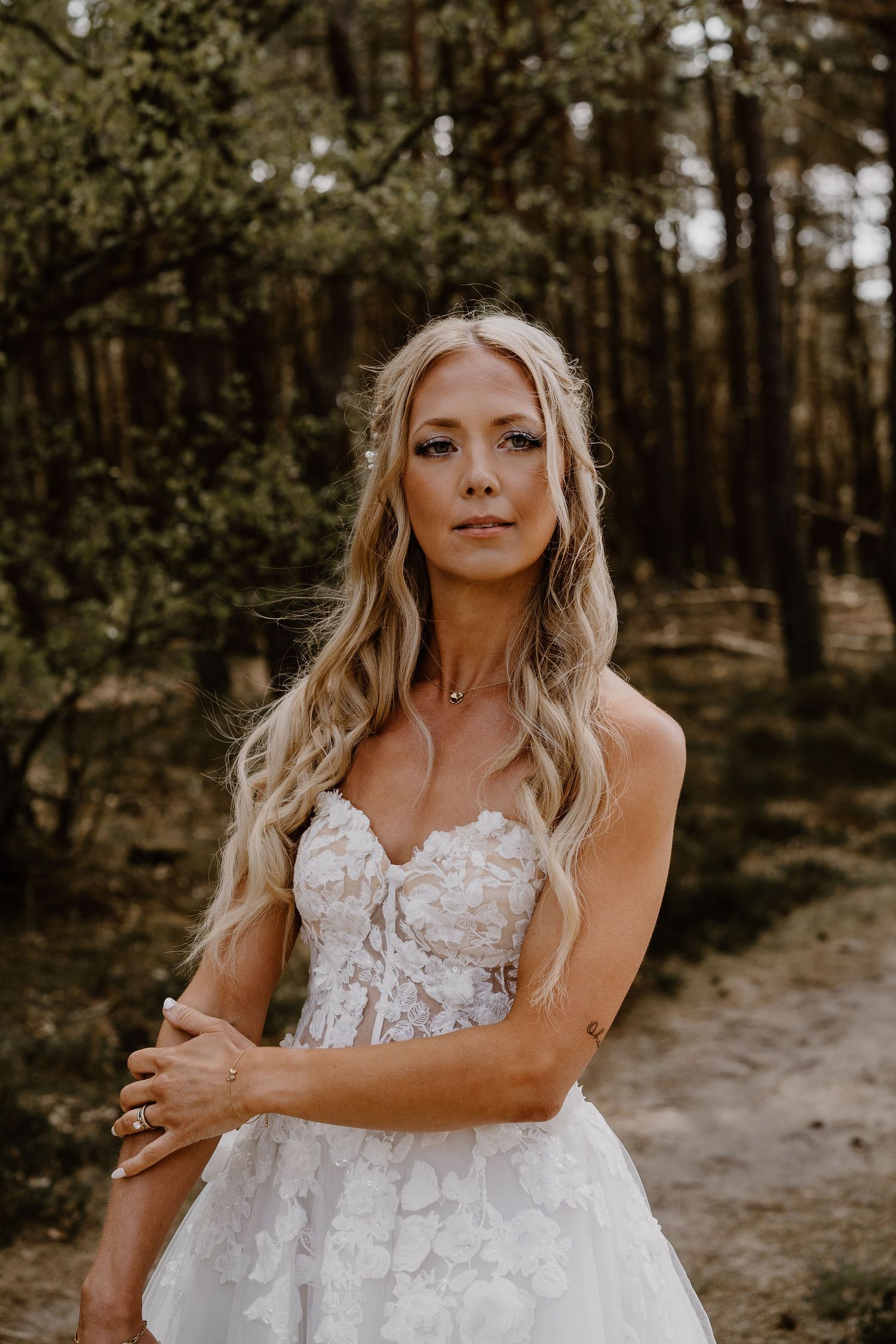 A bride in a strapless lace wedding gown poses in a wooded forest setting with warm natural lighting.