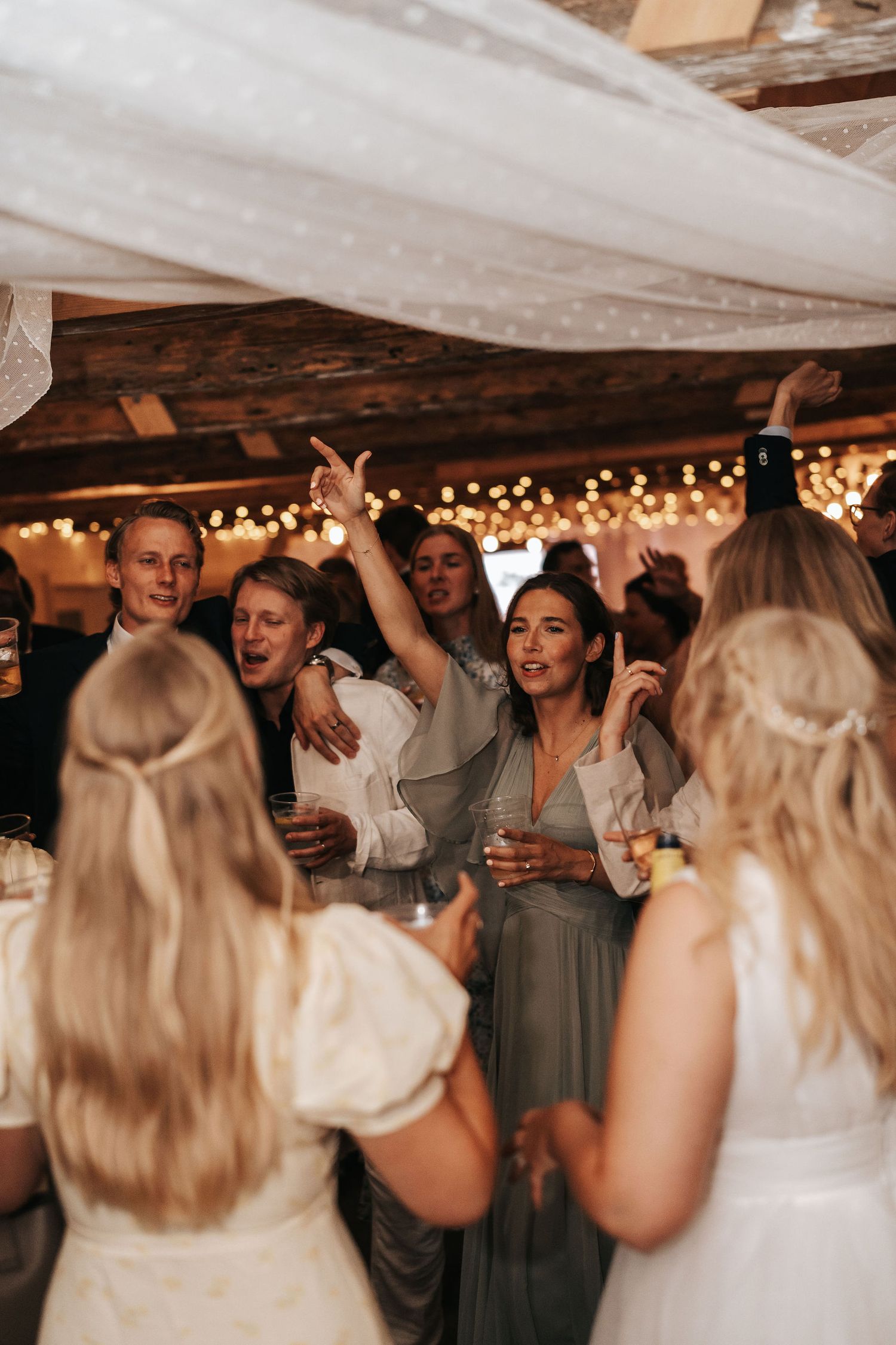 A group of wedding guests dancing and celebrating together under string lights at an indoor reception venue.