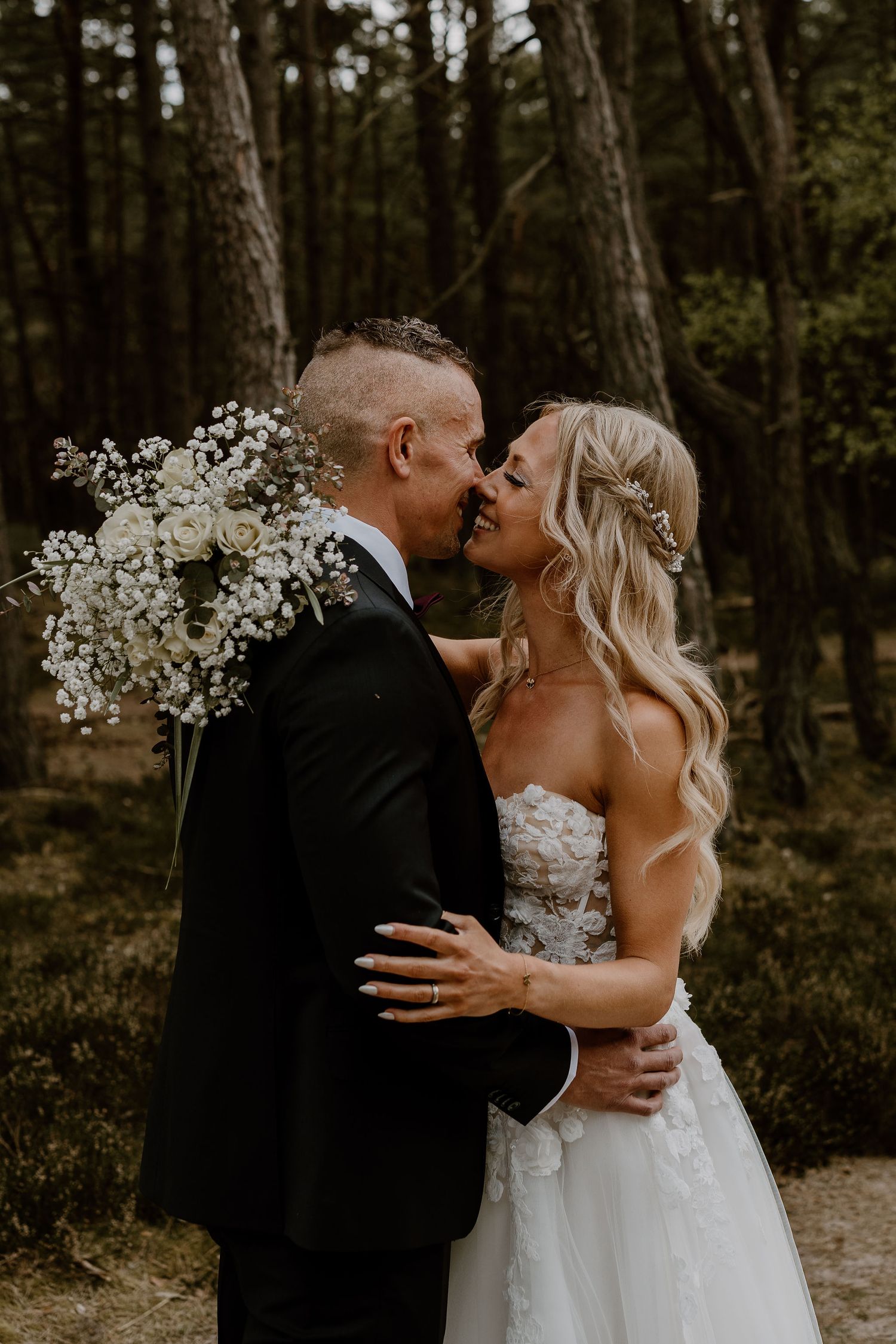 A couple shares a romantic kiss in a forest setting while the bride holds a white baby's breath bouquet.
