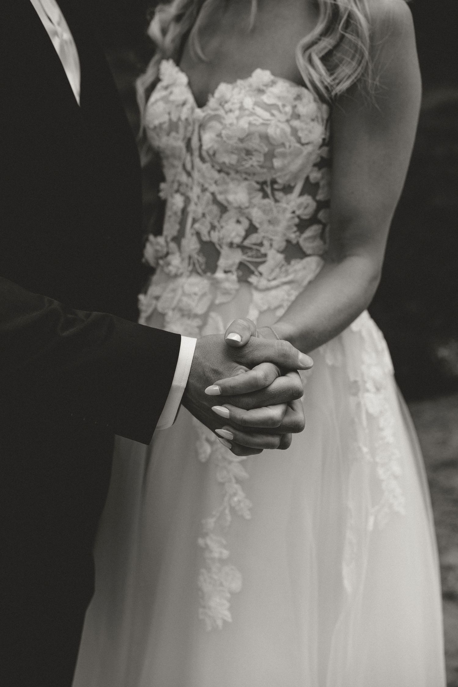Close-up black and white photo of a couple holding hands, showing detailed lace wedding dress bodice and suit sleeve.