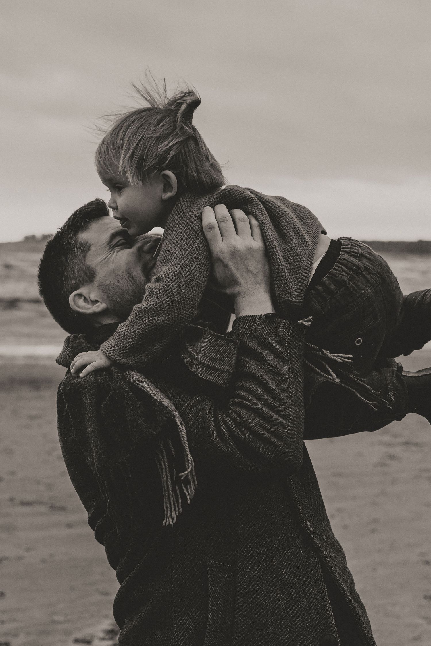 A tender black and white moment captured on a beach as parent lifts child in warm embrace against cloudy sky.