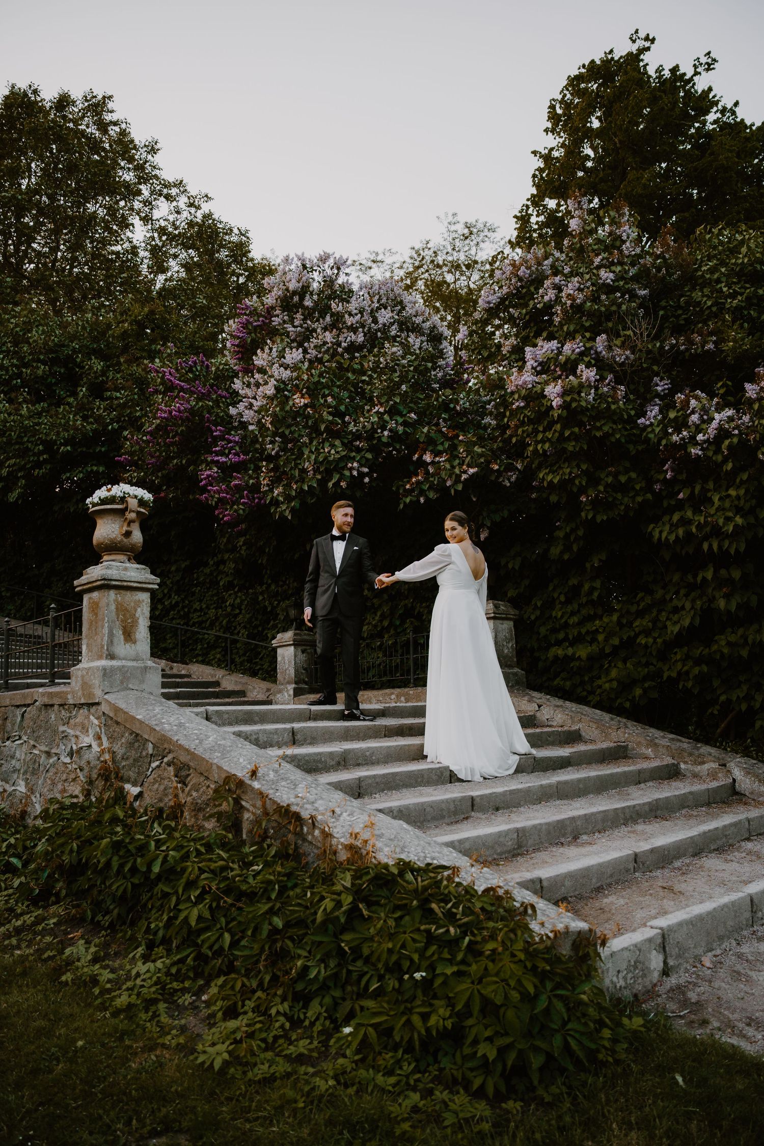 Elegant couple stands on vintage stone garden steps surrounded by blooming trees and greenery in romantic setting.