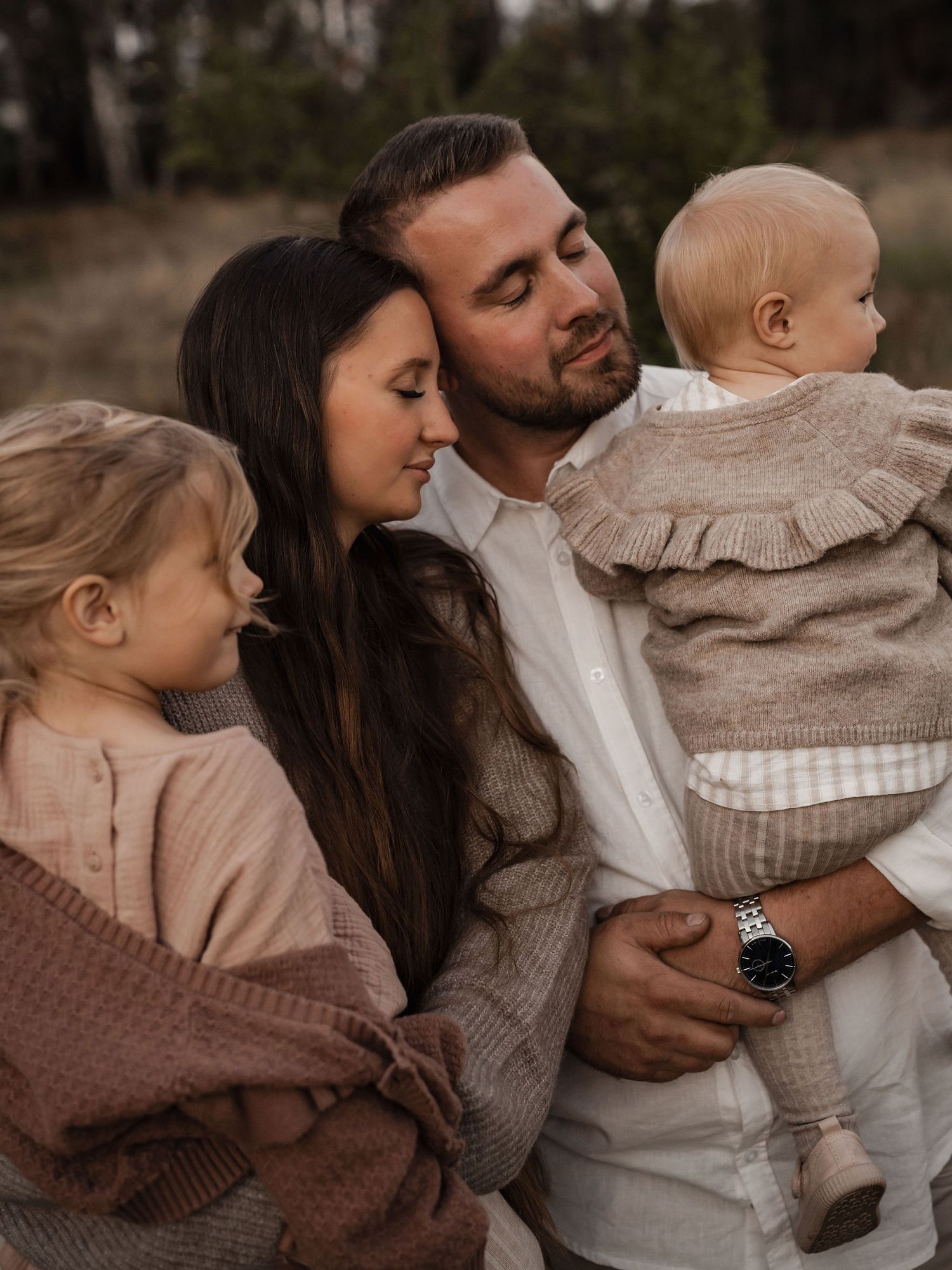 A family of four wearing neutral tones embraces while posing for an outdoor portrait in autumn.