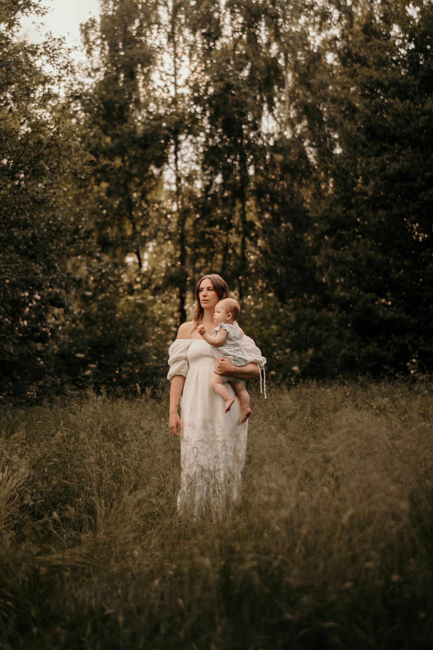 A person in a flowing white dress stands in a meadow at dusk surrounded by tall trees and wild grass.