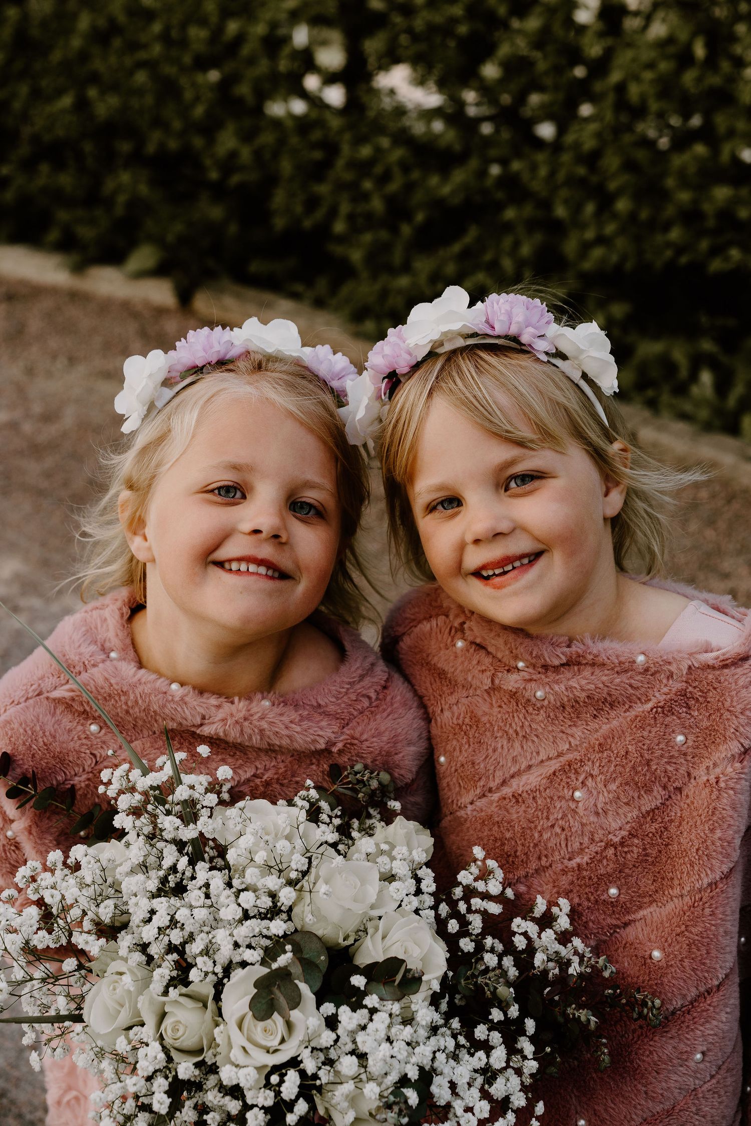 Two young attendants in pink wraps hold white floral bouquets and wear flower crowns during a wedding celebration.
