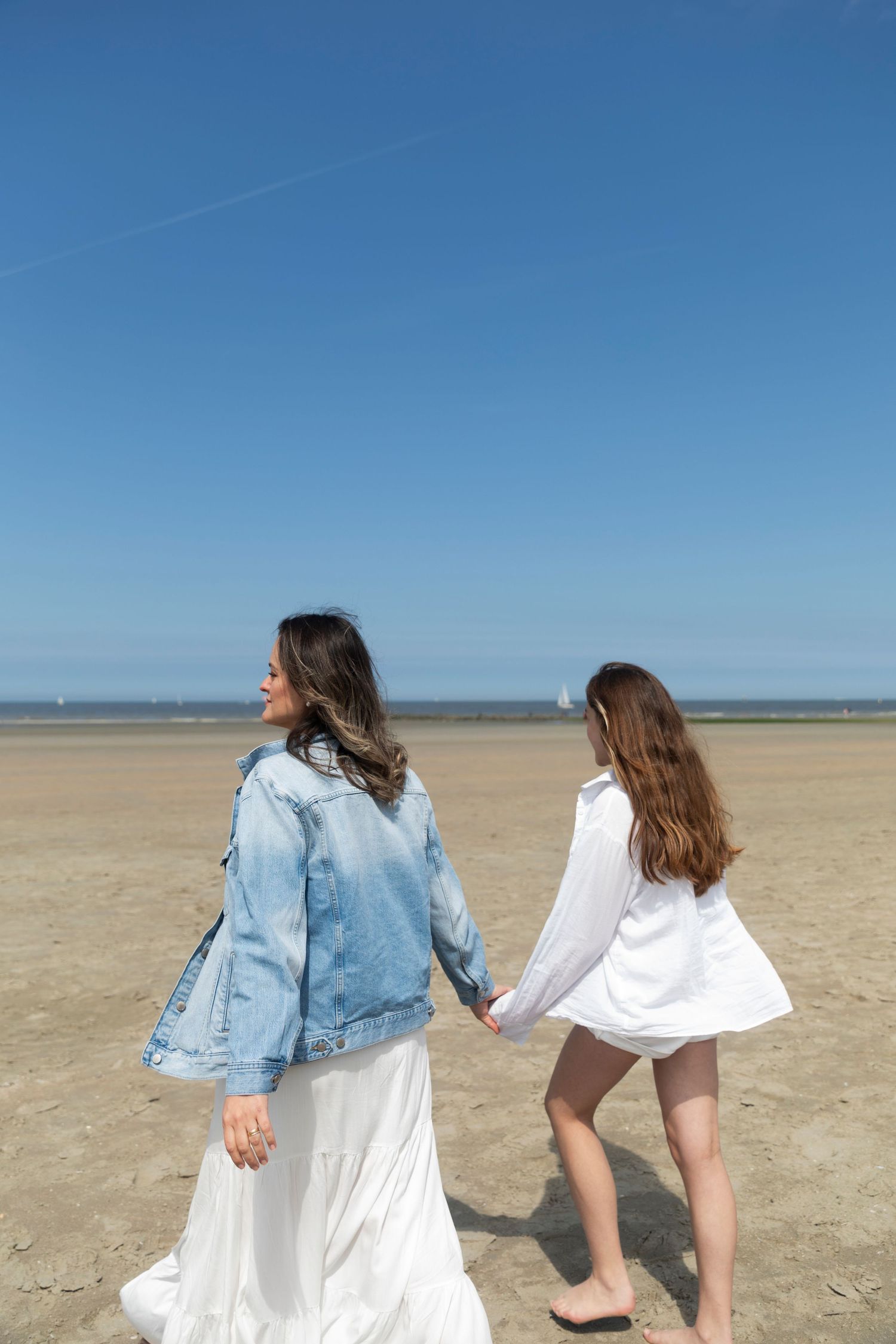 Two women holding hands walking on a sandy beach under a clear blue sky, one wearing a denim jacket and white skirt.