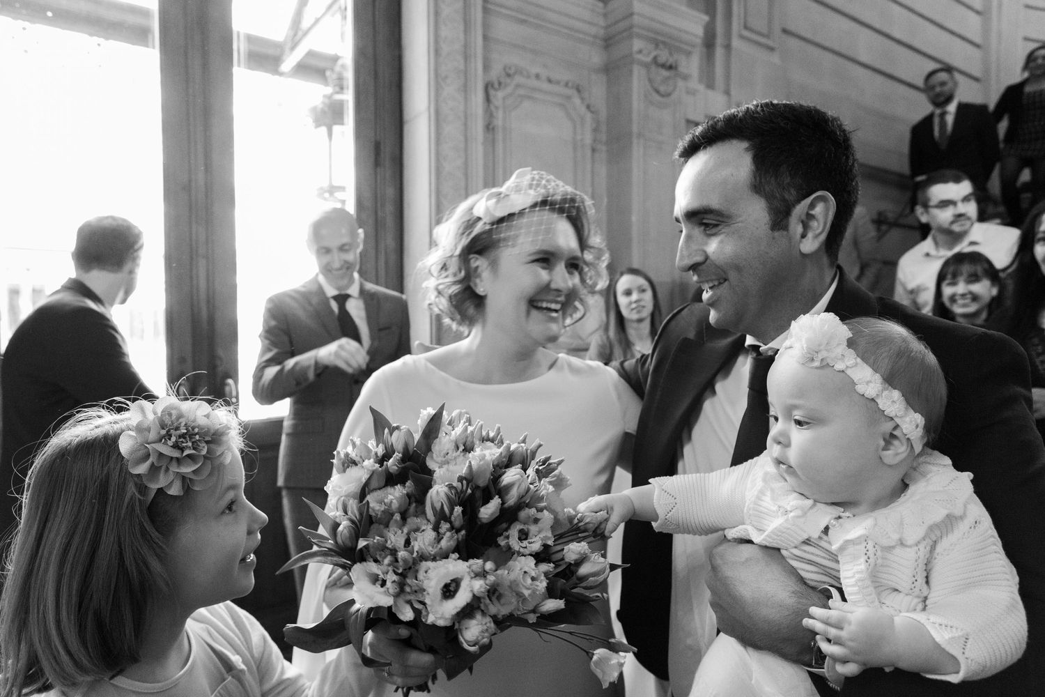 Black and white wedding photo of smiling bride with bouquet and groom holding baby, surrounded by guests at elegant venue.