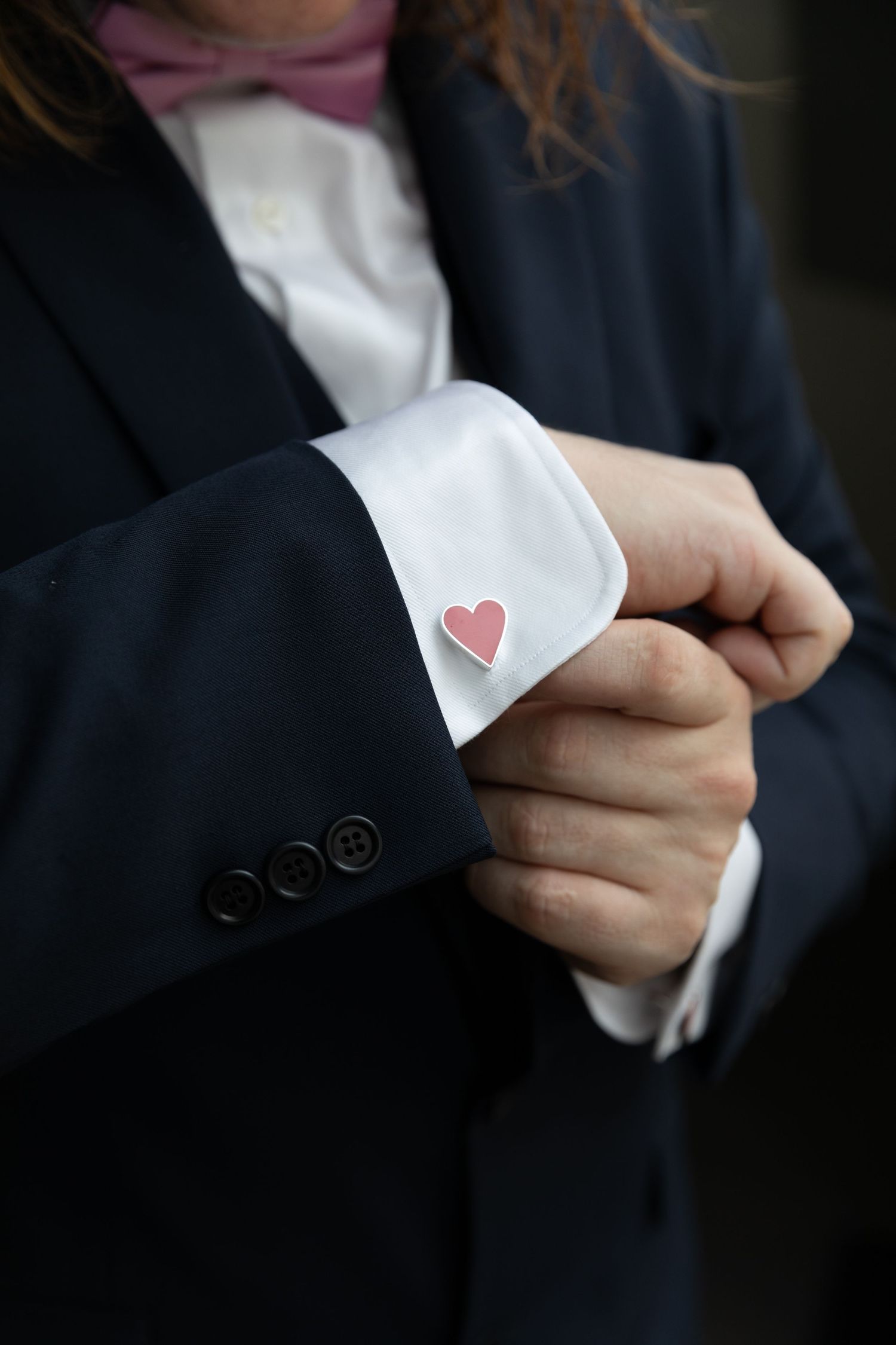 Person in dark suit adjusting heart-shaped pink cufflink on white dress shirt sleeve with pink bow tie.