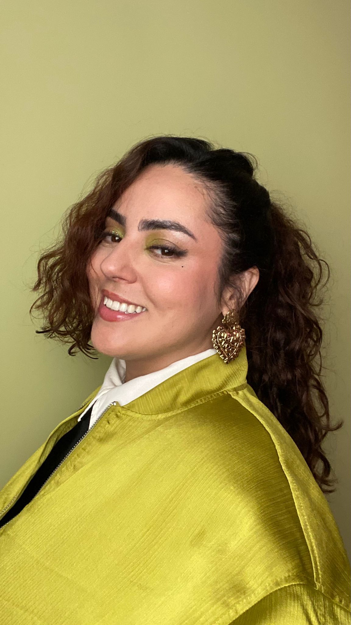 Smiling woman with curly hair wearing a chartreuse jacket and gold heart earrings against a green background.