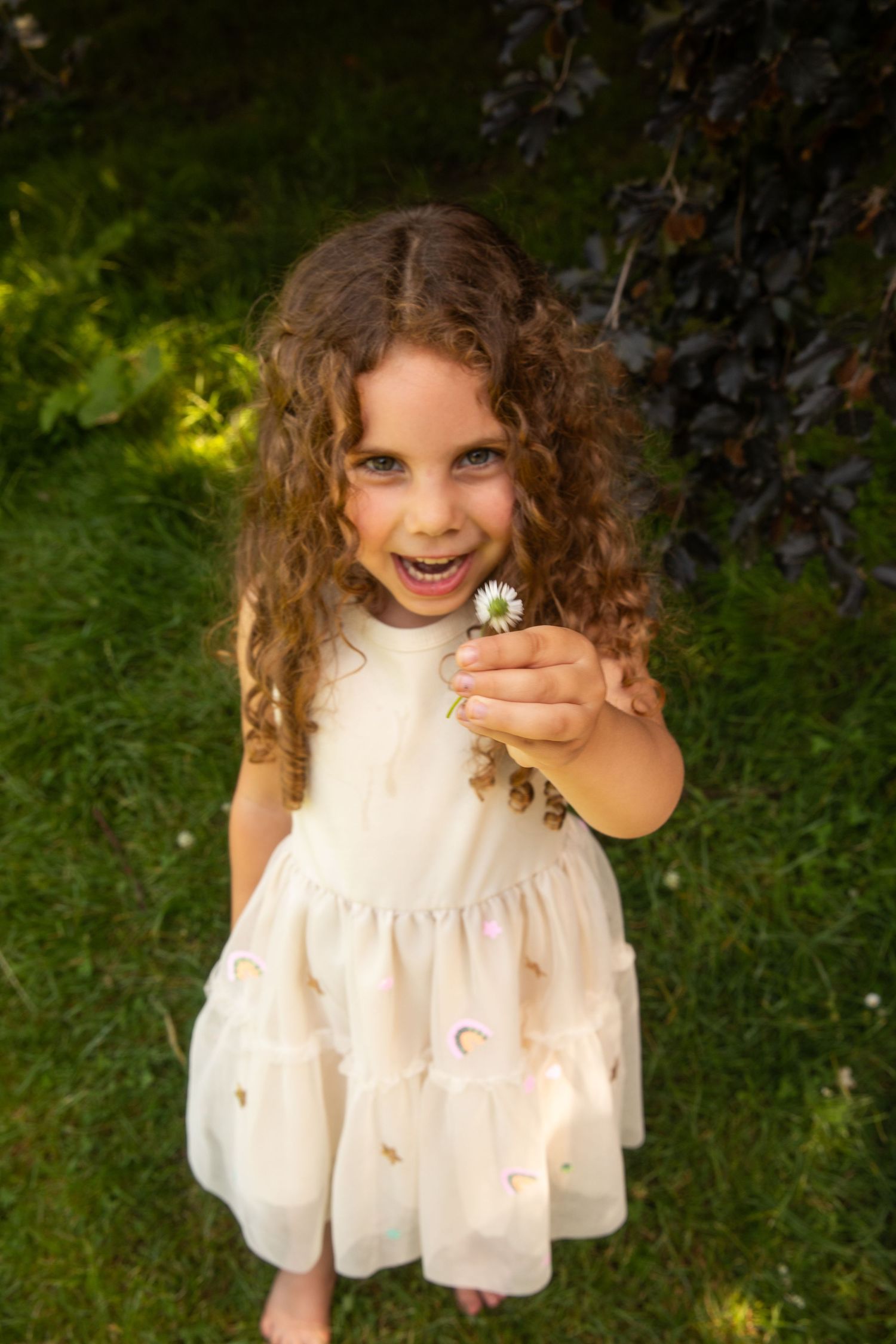 A joyful curly-haired girl in a white dress holds a daisy flower outdoors in a lush green garden.