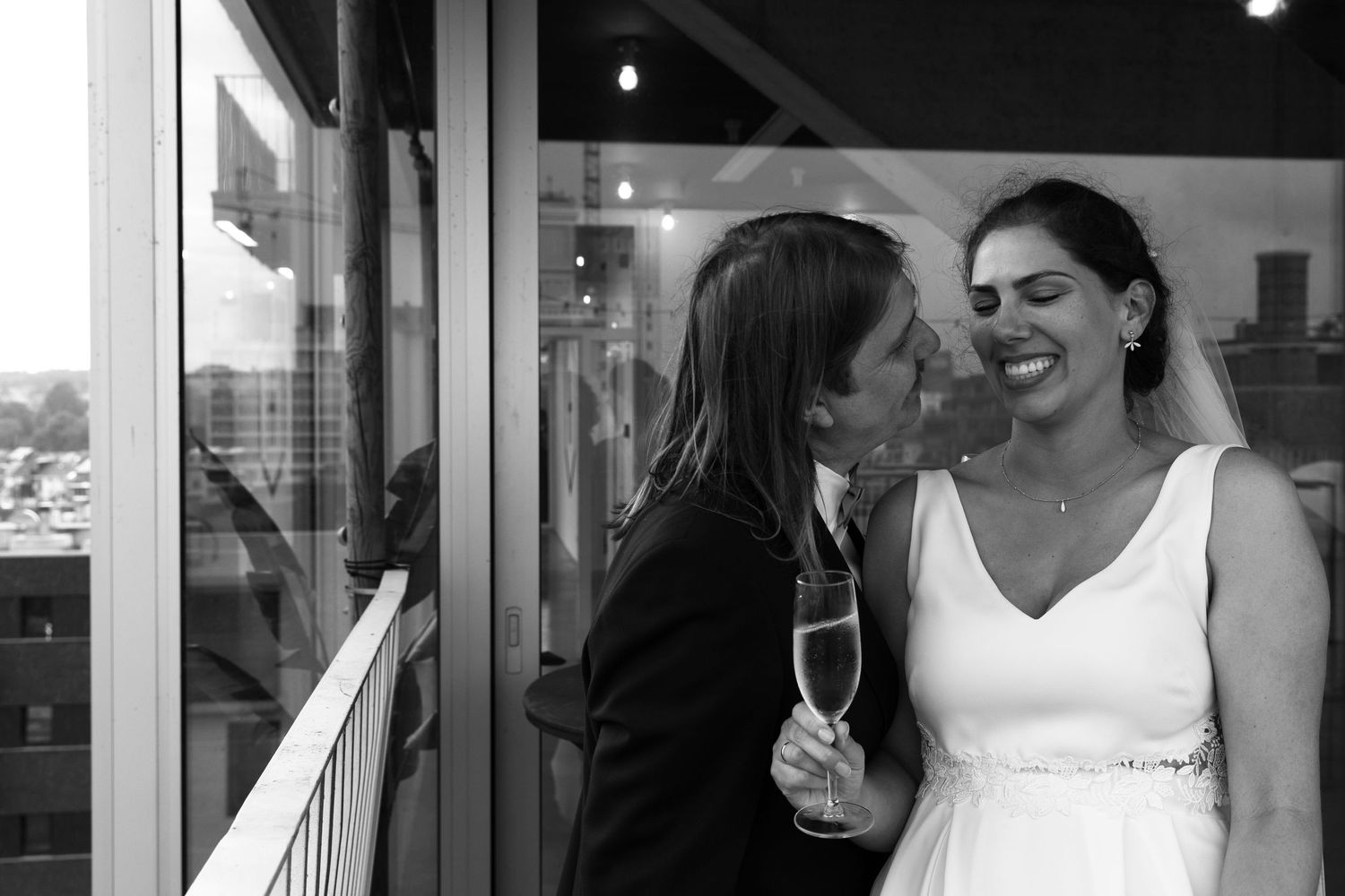 Black and white photo of a bride holding champagne being kissed on the cheek at an outdoor wedding reception.