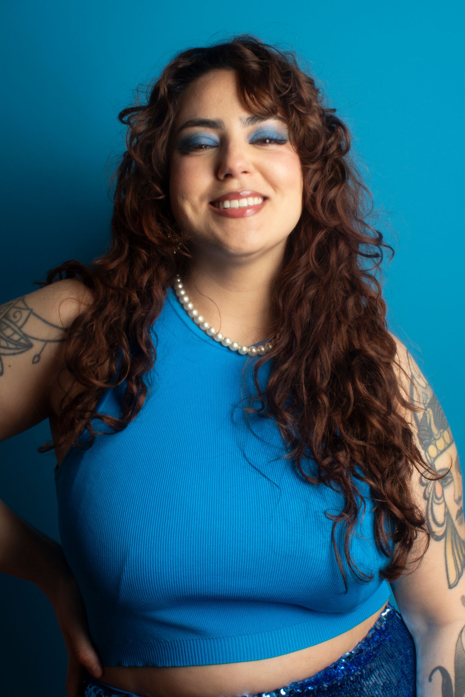 Smiling woman with curly auburn hair, blue eyeshadow, pearl necklace, and tattoos wearing a blue crop top.